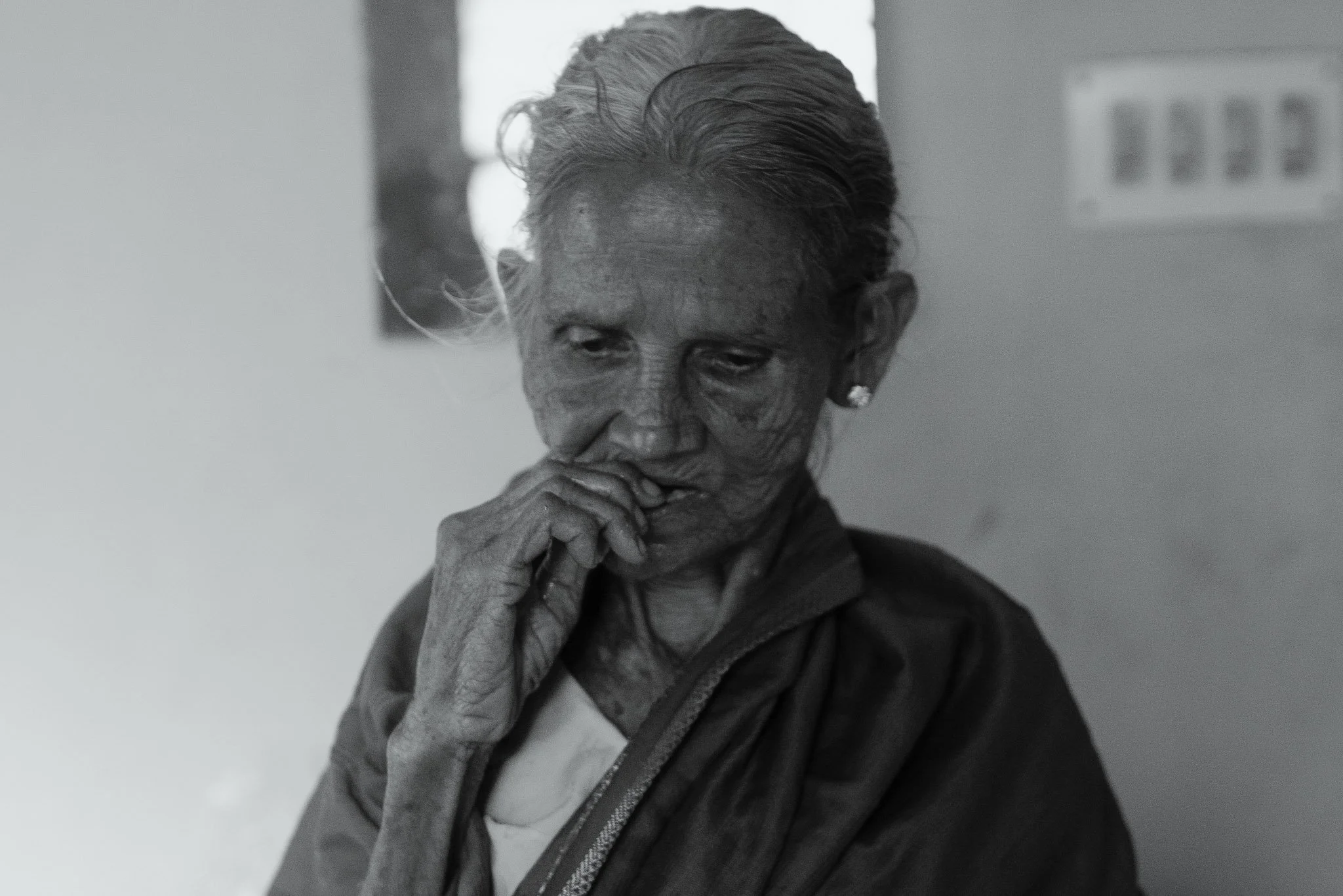 A black and white photo of an elderly woman with short hair, looking down with her hand near her mouth.