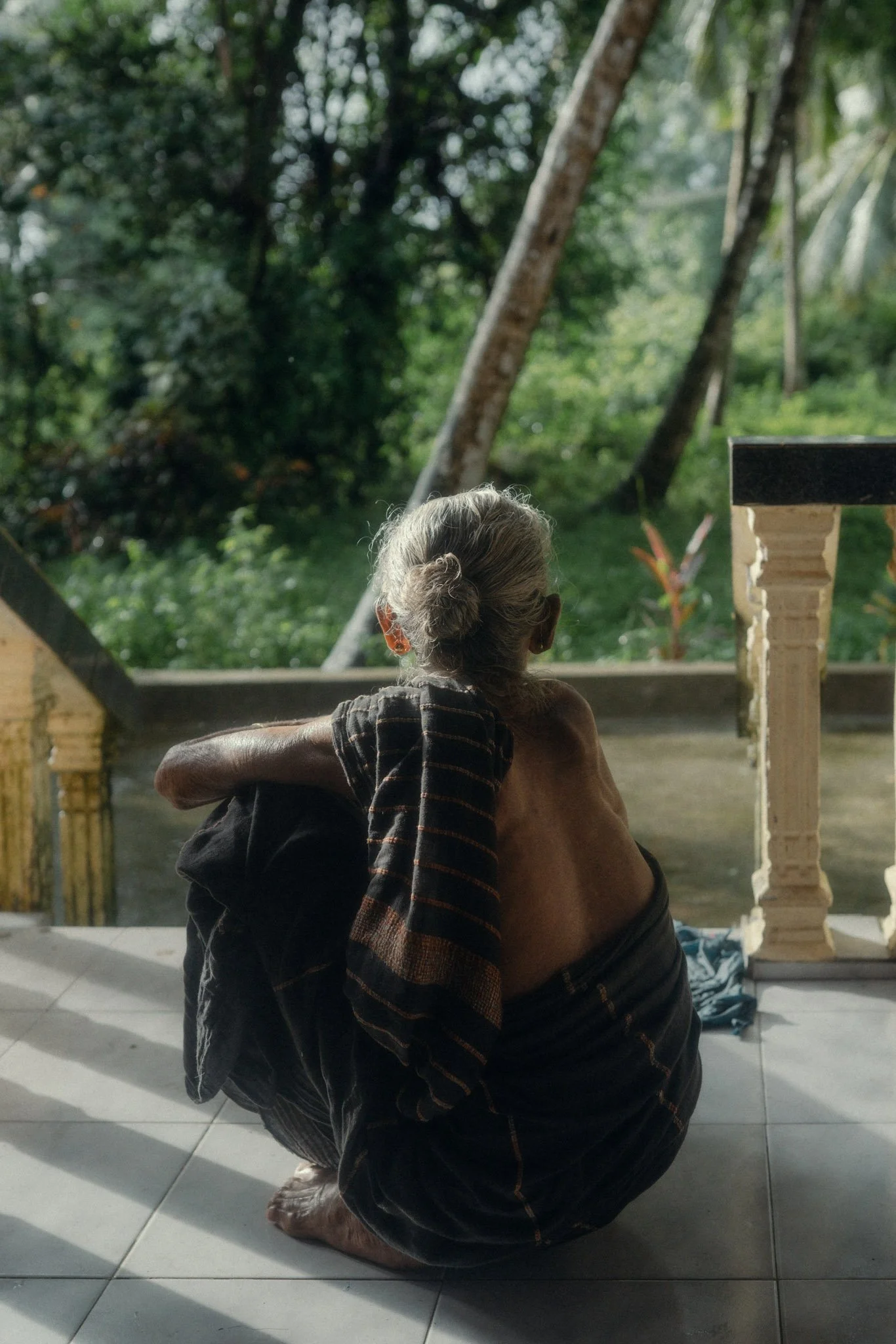 Older woman with gray hair sitting outdoors on a tiled floor, looking towards lush green trees, with a wooden railing in the foreground.