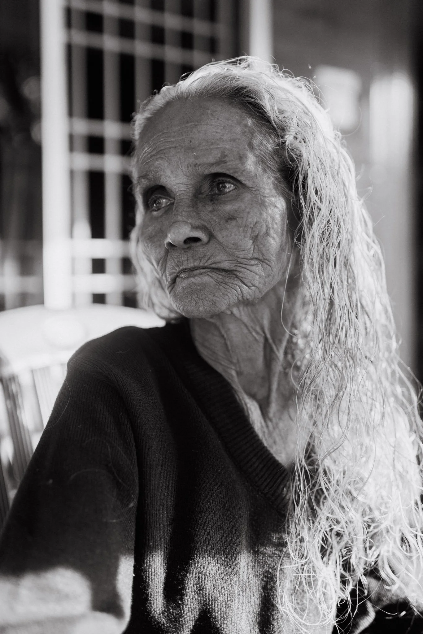 Black and white photo of an elderly woman with long, wavy white hair. She is looking upwards with a contemplative expression. The background shows a window with bars.