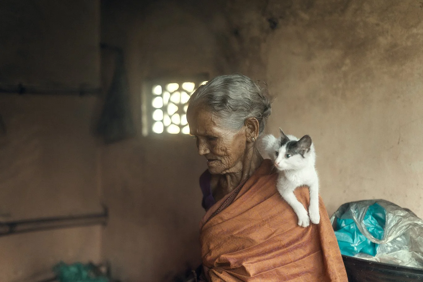 An elderly woman with gray hair in a bun, wearing traditional orange clothing, standing inside a rustic room with rough walls and small window. A white and gray cat rests on her shoulder, looking to the right.
