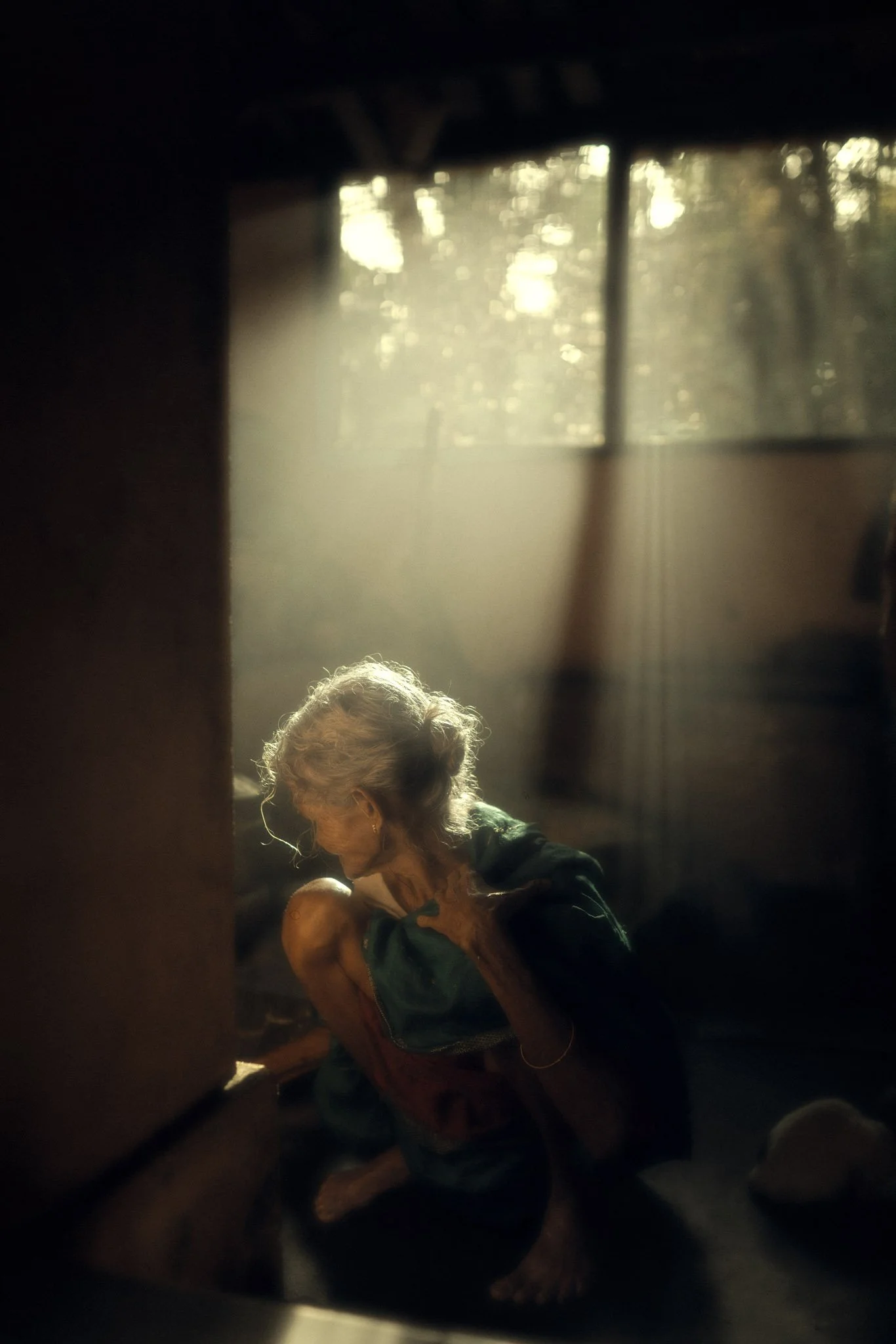 An elderly woman with white hair, wearing a sleeveless top, sitting in a dimly lit room with sunlight filtering through a window with curtains, creating a warm glow.