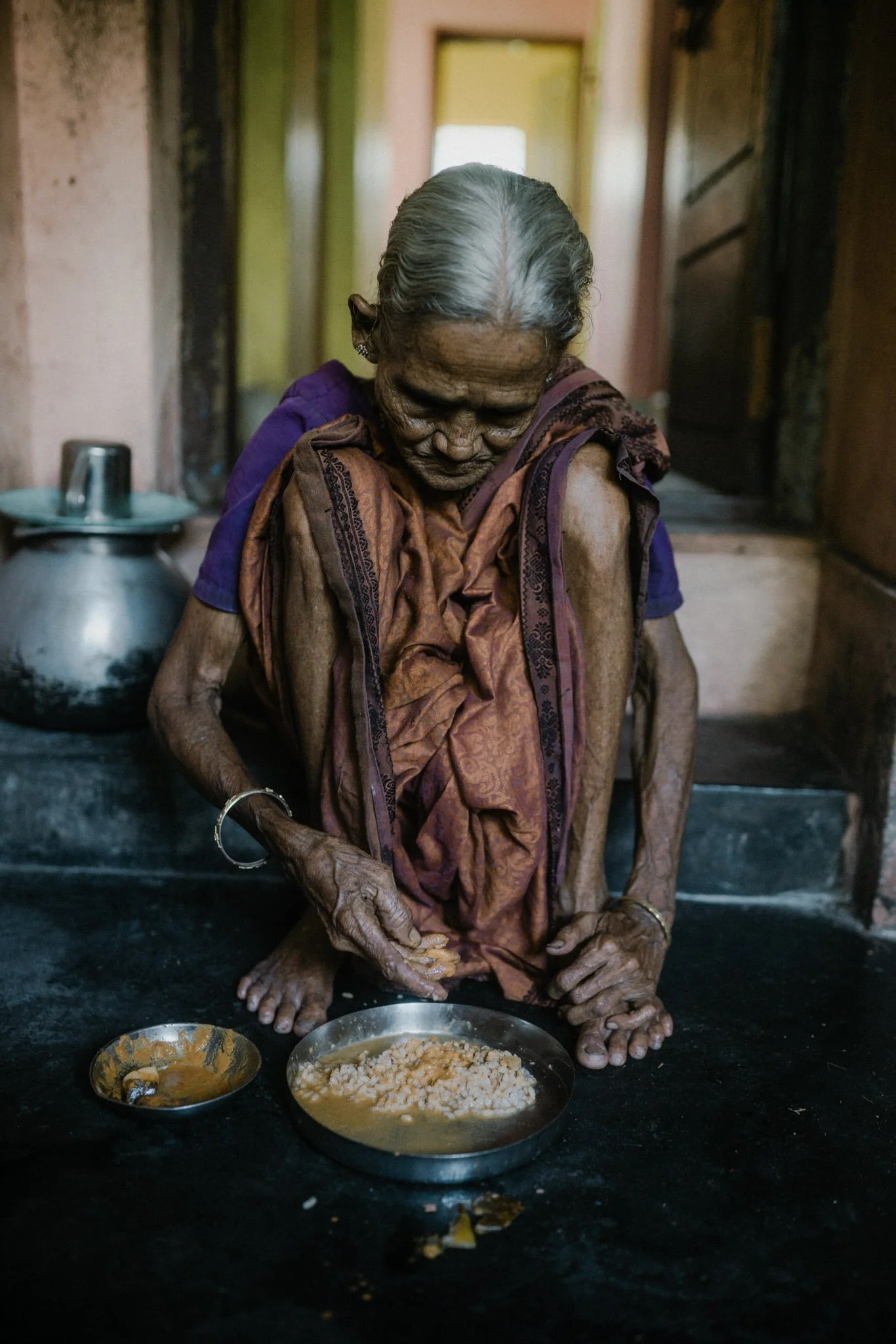 An elderly woman with gray hair and dark skin, wearing a traditional sari, is sitting on the floor of a modest kitchen, preparing food. There are two small bowls in front of her with food ingredients. The background shows an open doorway and kitchen 