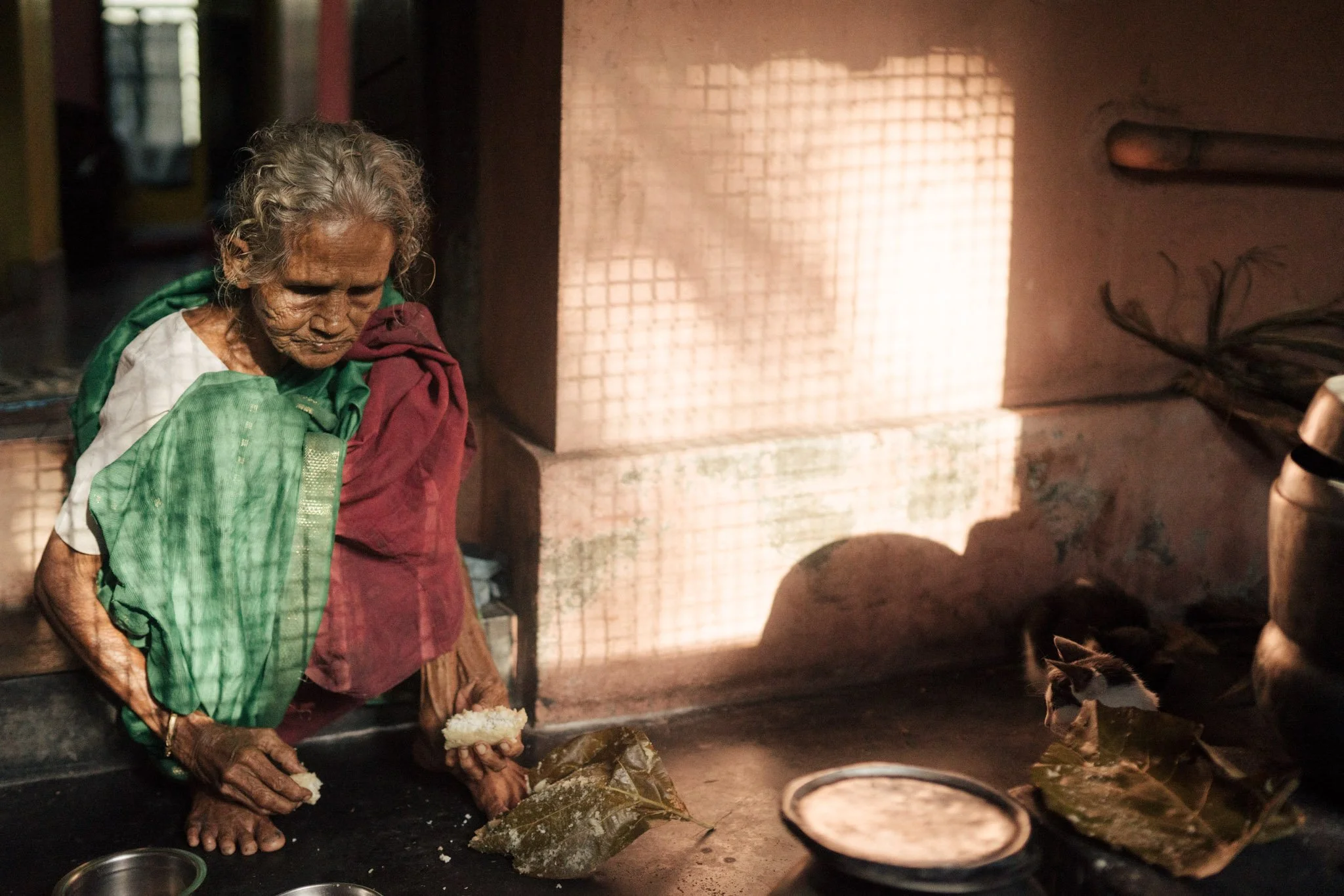 An elderly woman with gray hair and dark skin, wearing a green and maroon saree, is kneeling and preparing food in a rustic kitchen. She is holding a piece of rice or bread and has a slight smile. The background shows a brick wall and sunlight castin