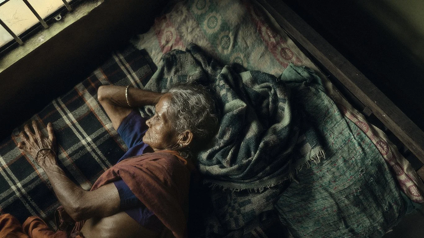 An elderly woman with gray hair sleeping on her side on a bed with patterned bedsheets and blankets, wearing a blue shirt and a reddish shawl.