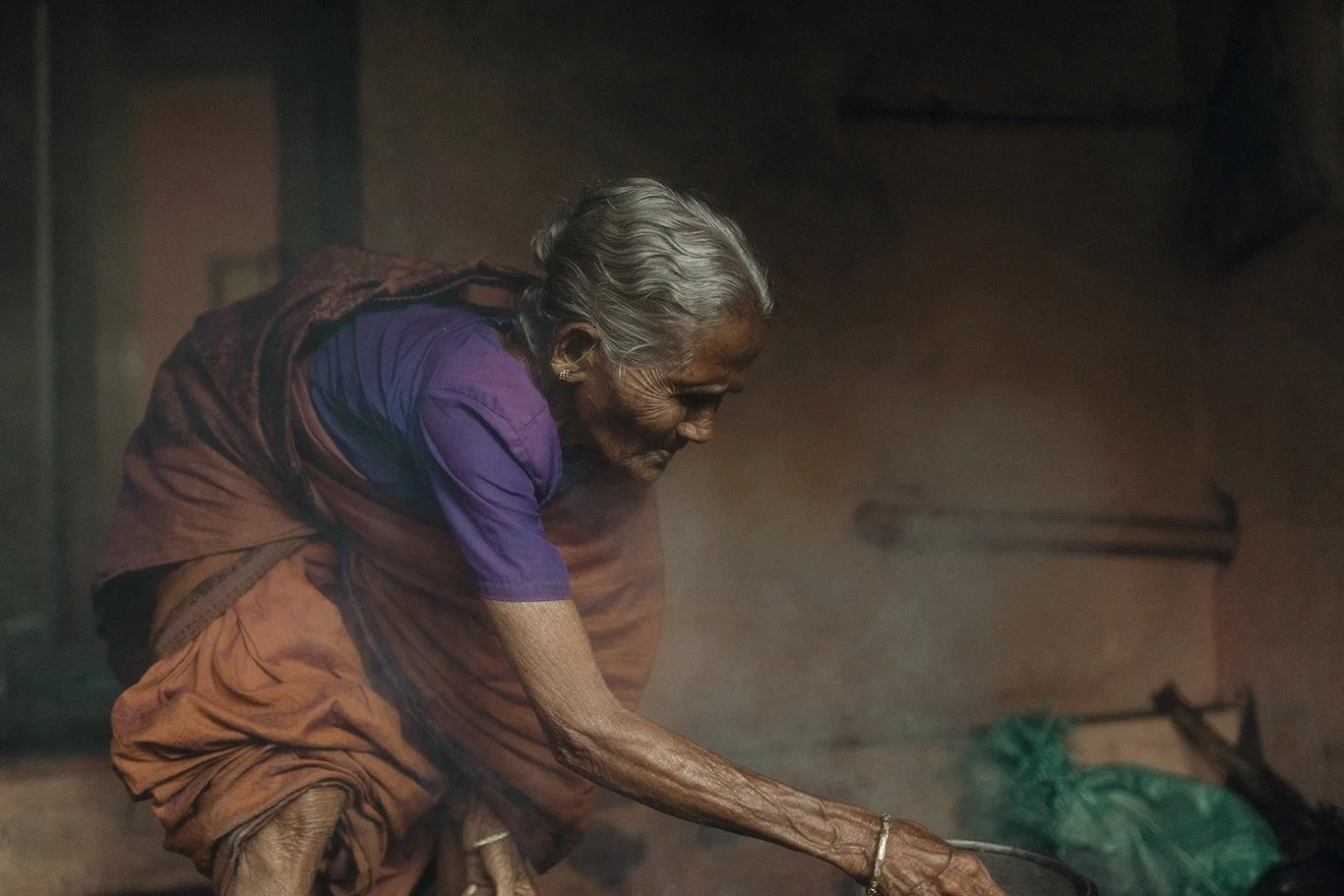 An elderly woman with gray hair, wearing a purple blouse and orange sari, cooking over a stove in a dimly lit room.