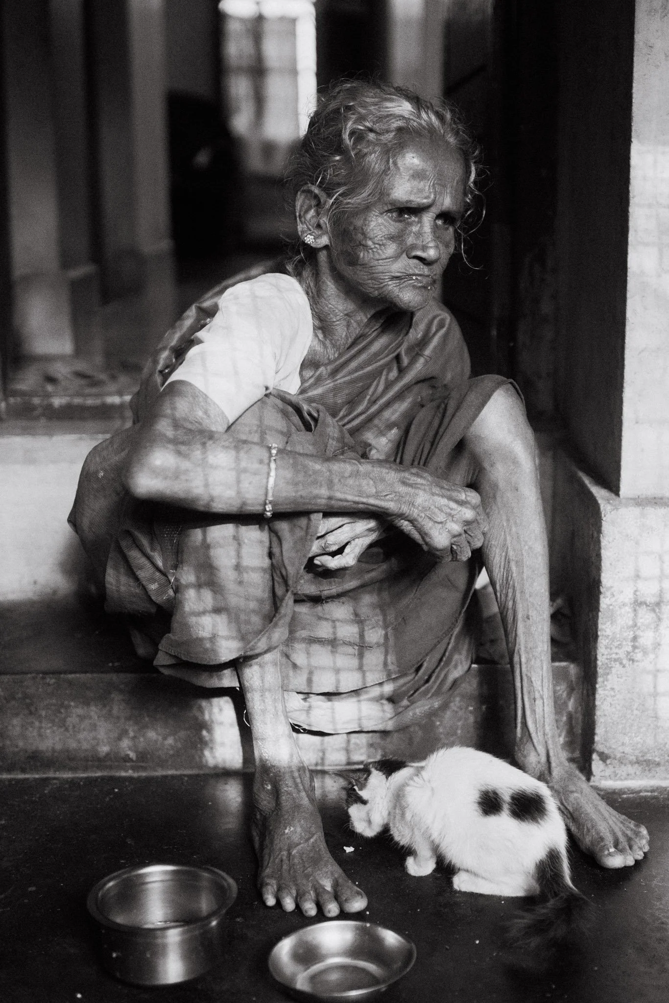 An elderly woman with wrinkled skin sitting on a small stool in a modest room, wearing traditional clothing and earrings. A calico kitten is nuzzling her leg near her feet. There are two empty metal bowls on the floor in front of her.