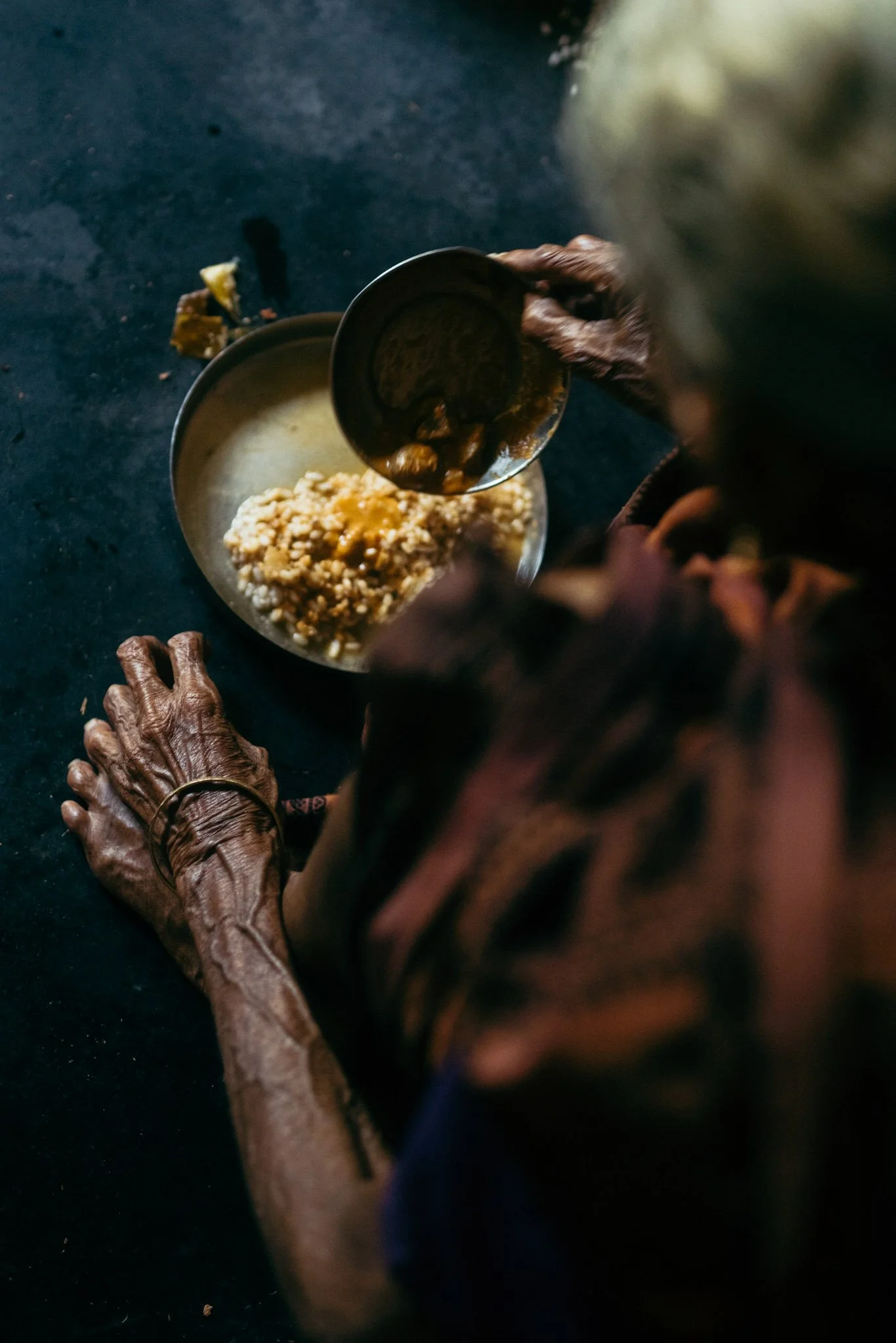 Close-up of elderly person's wrinkled hands reaching into a metal bowl full of food, with another container nearby and some food on the dark surface.