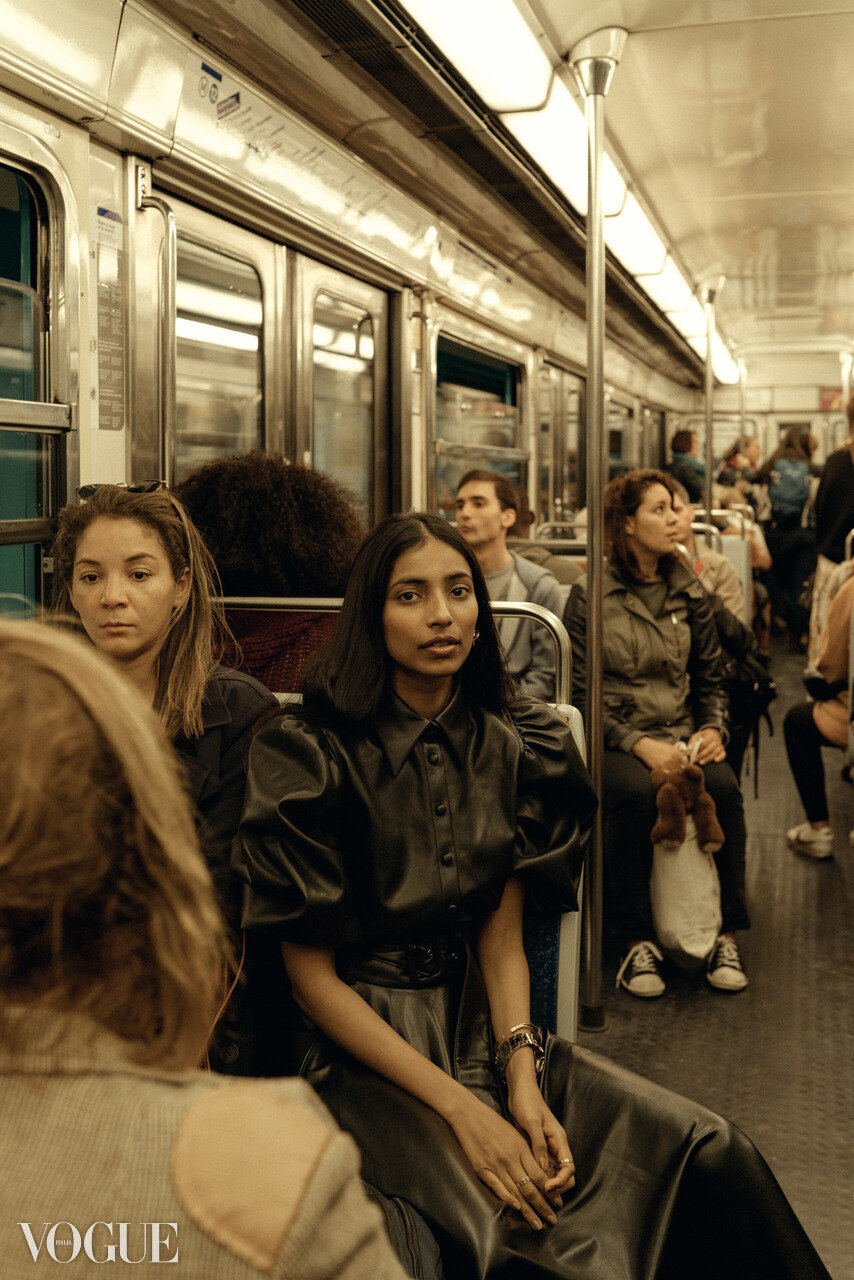 People sitting on a subway train, including a woman with black hair and black leather outfit, a woman with light brown hair, and a woman with a teddy bear, with others visible in the background.