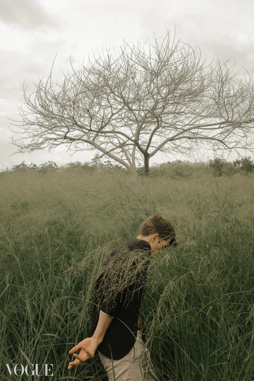 A person in black and khaki clothing standing in tall grass with a leafless tree and cloudy sky in the background, Vogue logo in bottom left corner.