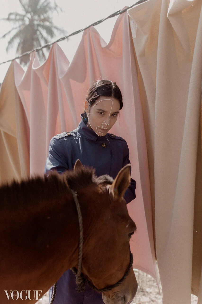 A woman with dark hair, wearing a navy jacket, stands behind a brown horse with a rope halter, against a backdrop of hanging pastel-colored fabrics and a palm tree, in an outdoor setting.
