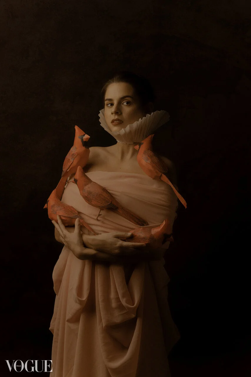 A woman in a beige dress with a ruffled collar is surrounded by orange birds resting on her arms and shoulders, against a dark background.