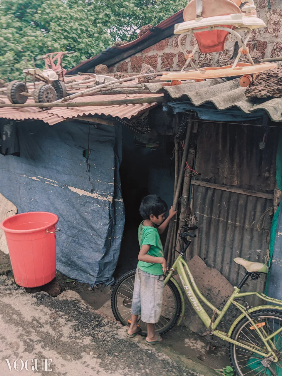 A young boy in a green shirt and shorts stands next to a yellow bicycle outside a makeshift home with corrugated metal walls and a tarp roof. He is holding onto the bicycle's handlebar and looking inside the home. There is a large orange plastic buck