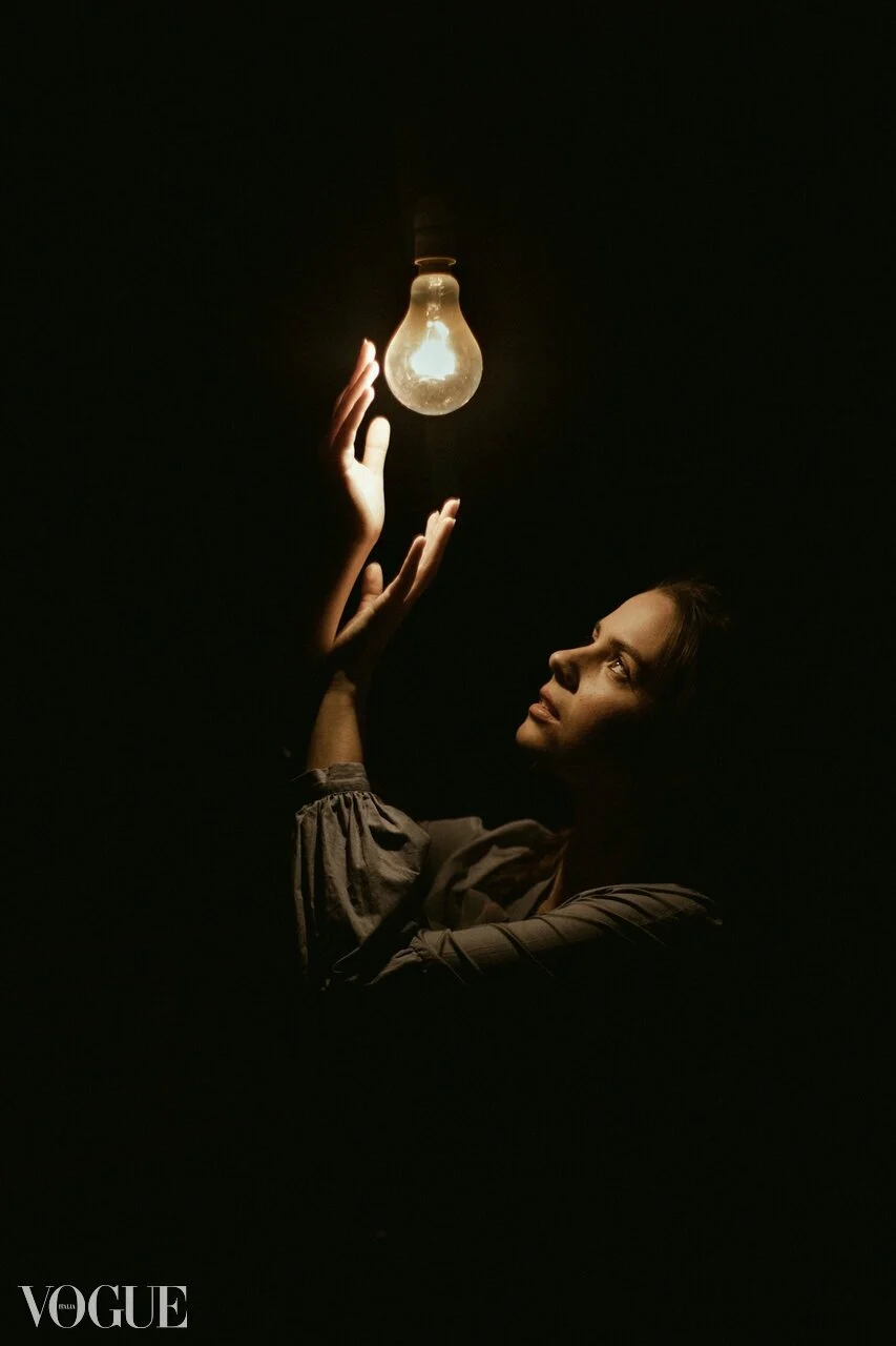 A woman in a dark environment is reaching up towards a glowing light bulb hanging from the ceiling.