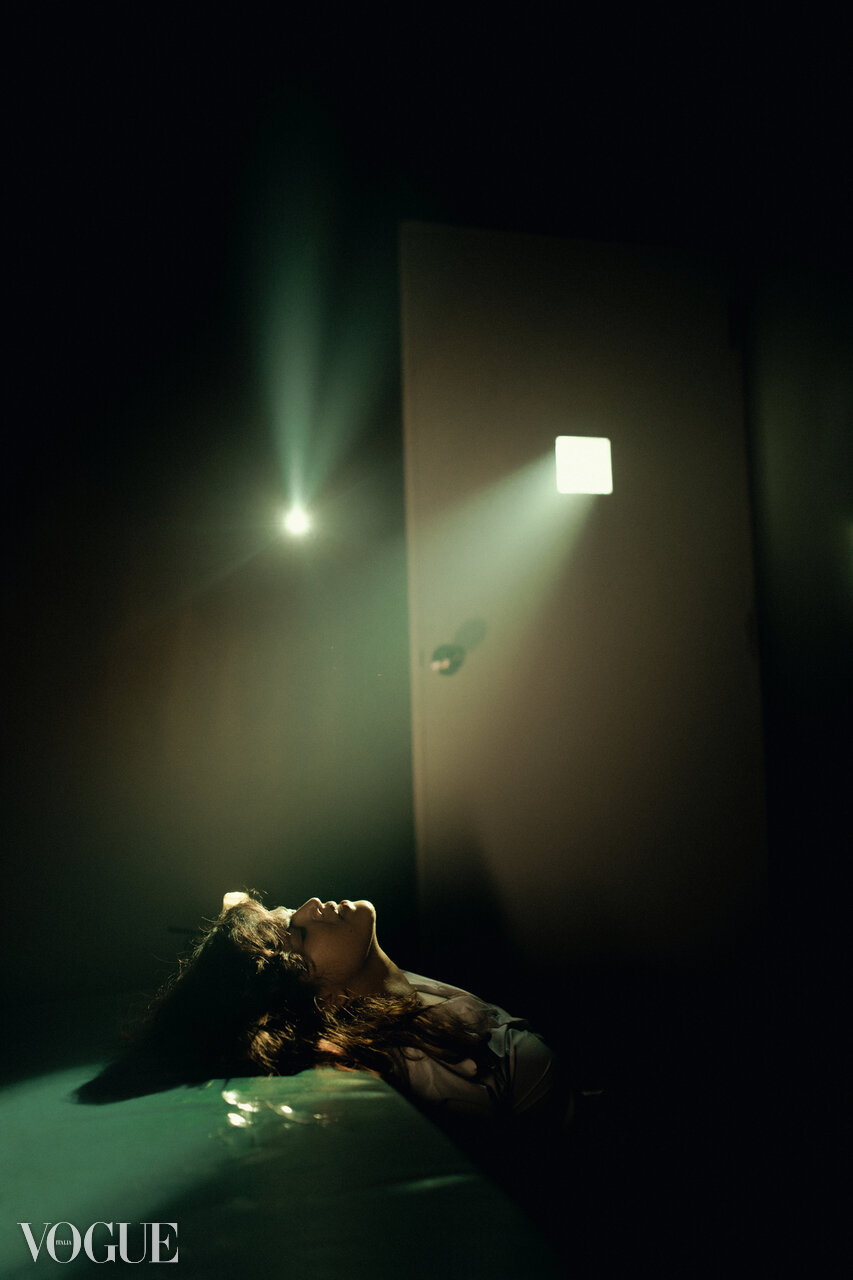 A woman lying on her back on the floor in a dark room with minimal lighting, illuminated by a bright light coming from a small square window in a closed door.