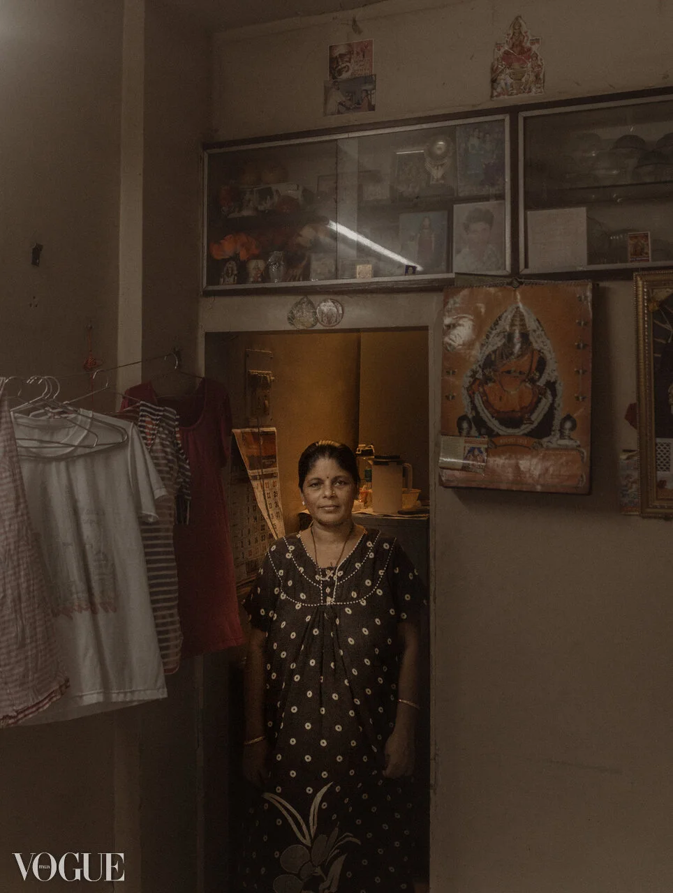 A woman stands in a doorway inside a small room decorated with religious images, clothes hanging on a rack, and framed pictures on the wall.