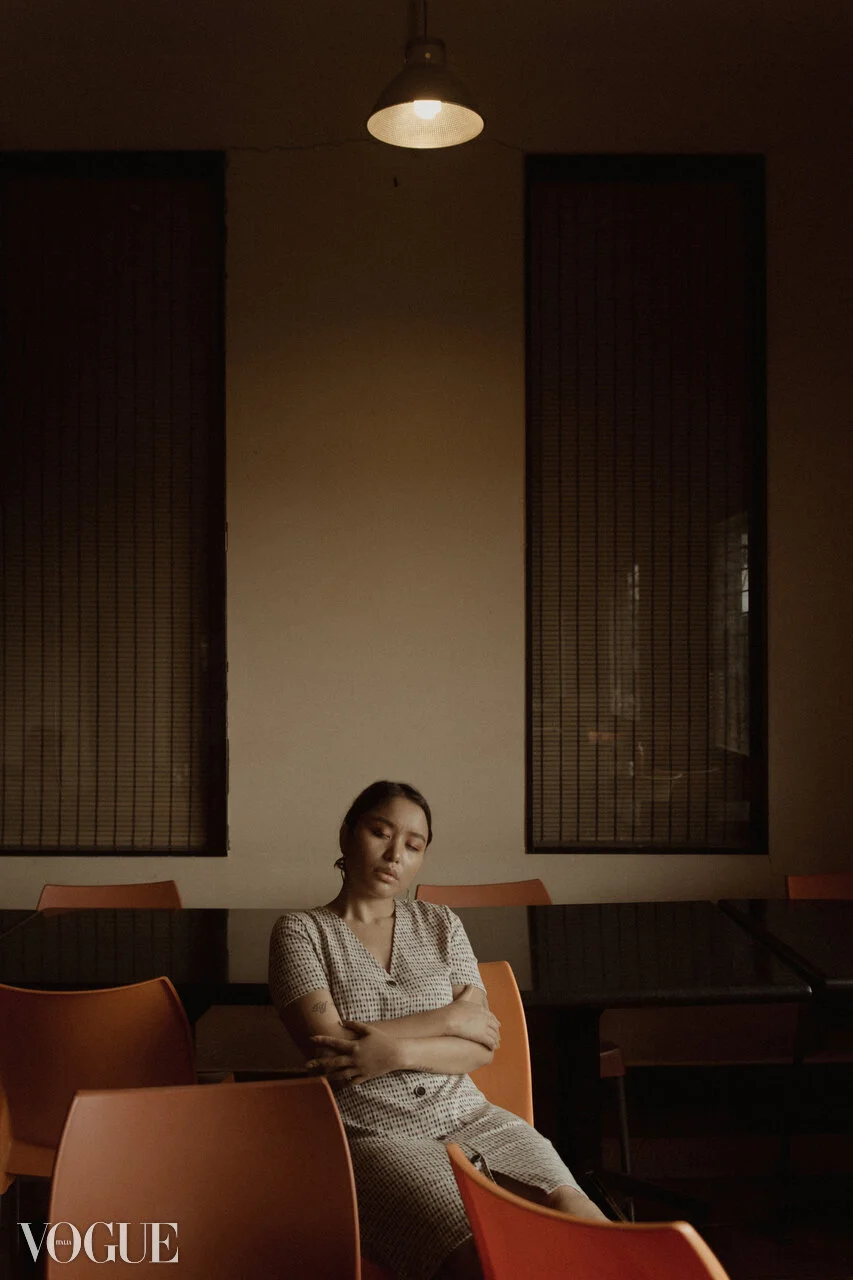 A woman sitting alone in a dimly lit room with orange chairs, a beige wall, and tall windows with blinds, wearing a checkered dress and looking down with arms crossed.