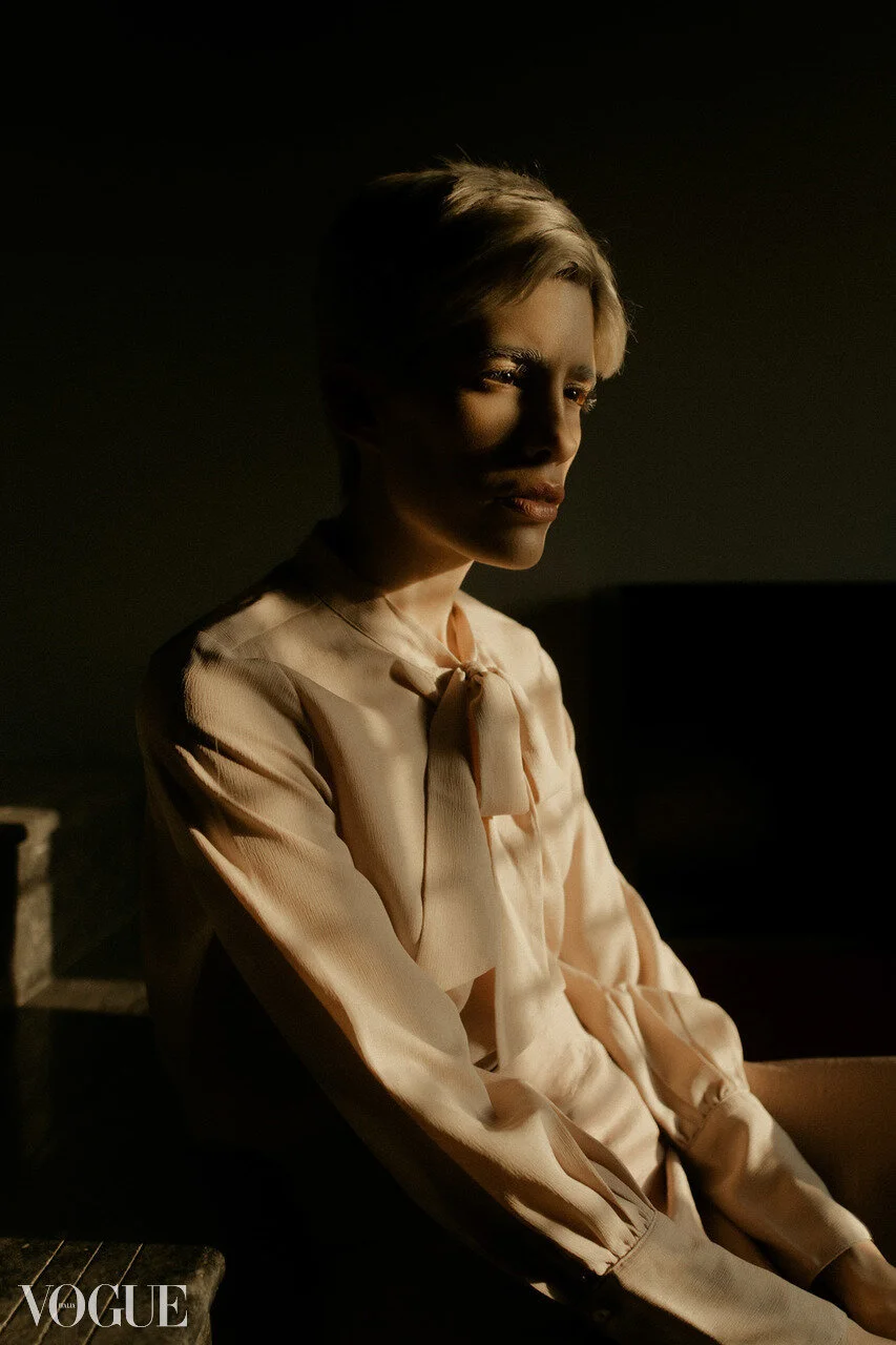 A woman with short blonde hair sitting in a dimly lit room, wearing a light-colored blouse with a bow at the collar, looking thoughtfully into the distance.