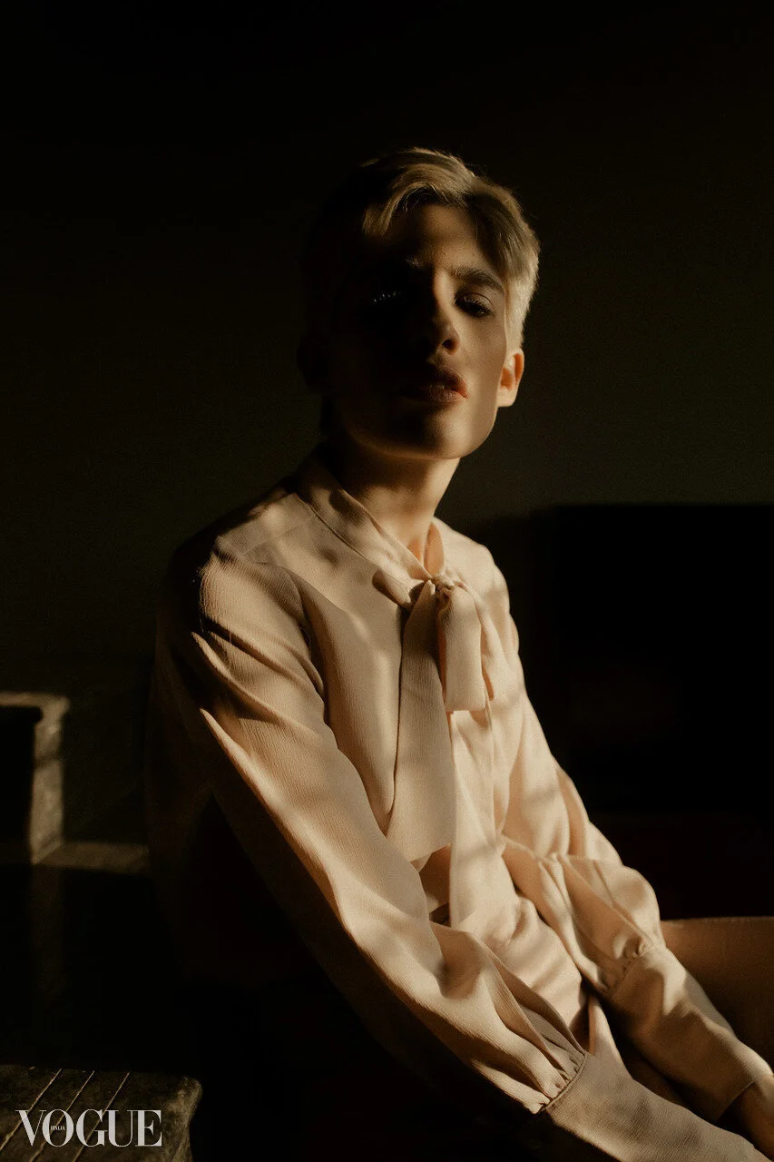 A woman with short blonde hair sitting in low lighting, wearing a light-colored blouse with a bow tie, looking towards the camera with a serious expression.