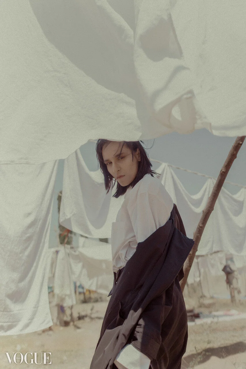 A woman with dark hair, wearing a white shirt and black jacket, standing outdoors among hanging white sheets and laundry lines, with a neutral expression.