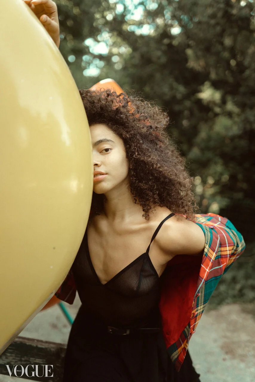 A young woman with curly hair poses outdoors behind a large yellow surfboard, wearing a black sheer top and a colorful plaid shirt draped over her shoulder.