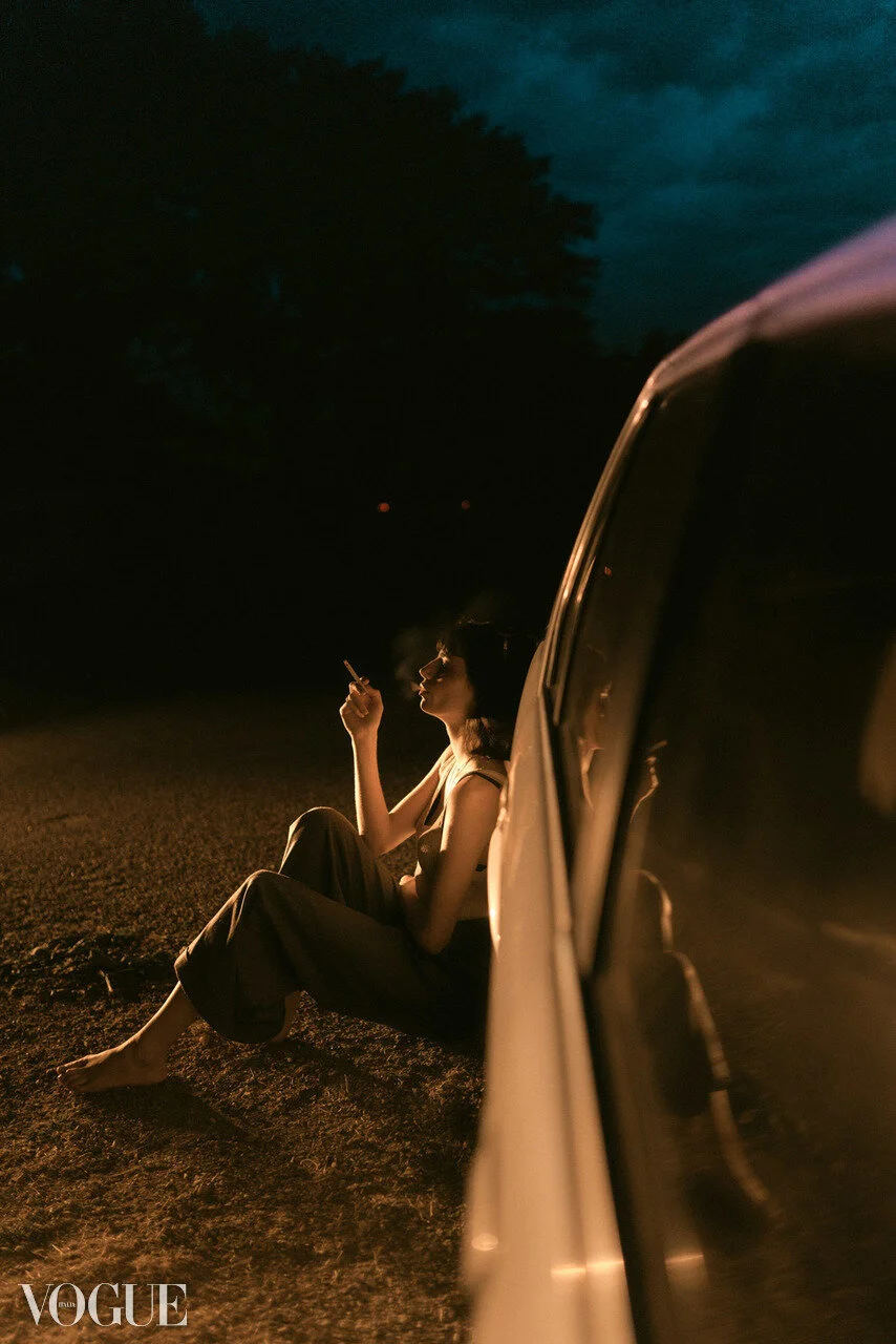A woman sitting on the ground outside at night, smoking a cigarette, with her reflection visible in the side mirror of a vehicle parked nearby.