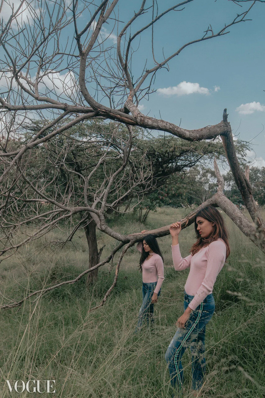 Two women standing in a grassy field with leafless trees, under a partly cloudy sky.