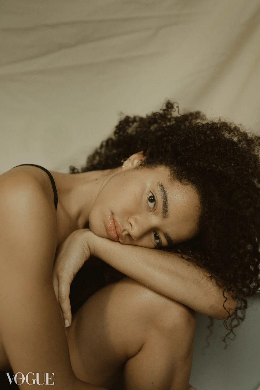 A young woman with curly hair resting her head on her knee and looking at the camera, lying against a neutral background, Vogue branding in the bottom left corner.