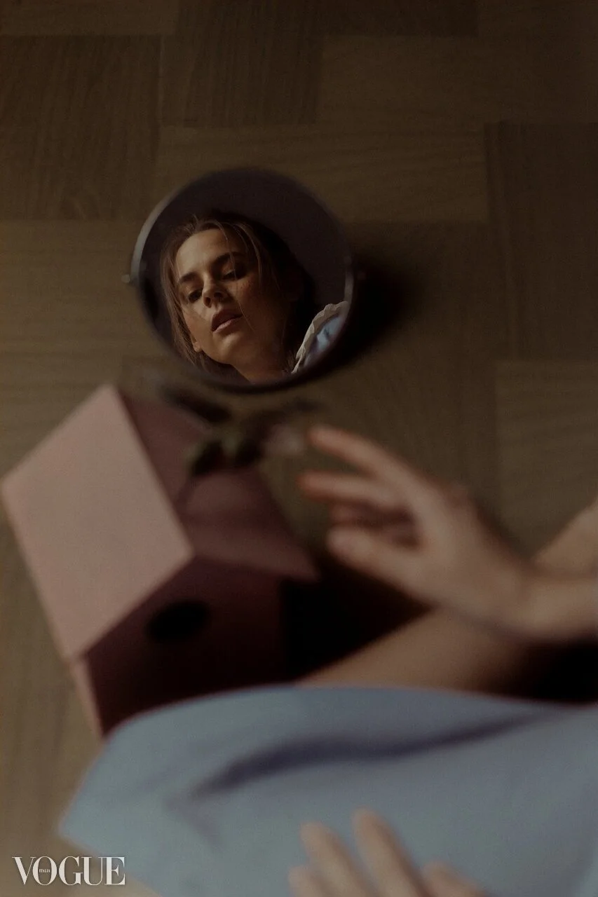 A woman taking a selfie, reflected in a round mirror on the floor, with a pink box and a hand visible in the frame, and a wooden floor background.
