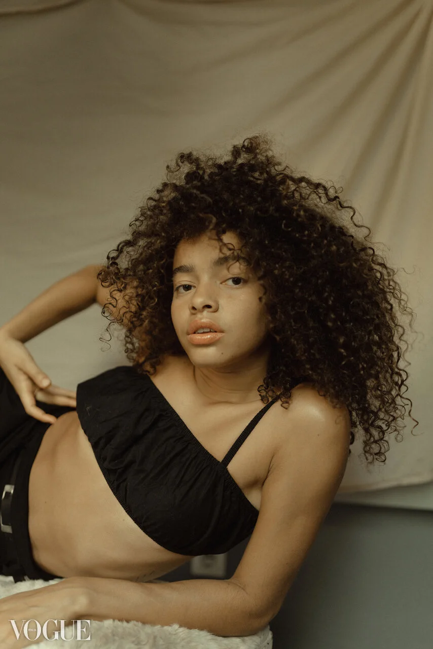 A woman with curly hair lying on her side on a beige rug, wearing a black top and shorts, with a beige backdrop.