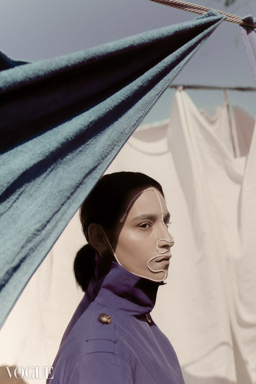 Fashion model with geometric face makeup and purple coat posing behind hanging laundry, Vogue logo in bottom left corner.