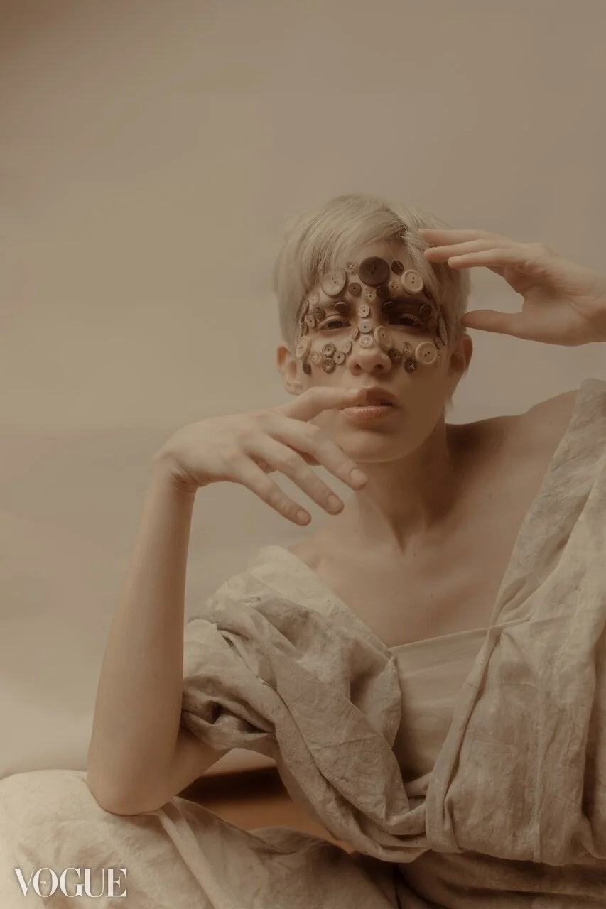 A person with short blond hair and no shirt, wearing multiple buttons on their face, posing with one hand near their forehead and the other near their chin, in a neutral room with beige walls, Vogue logo in the bottom left corner.