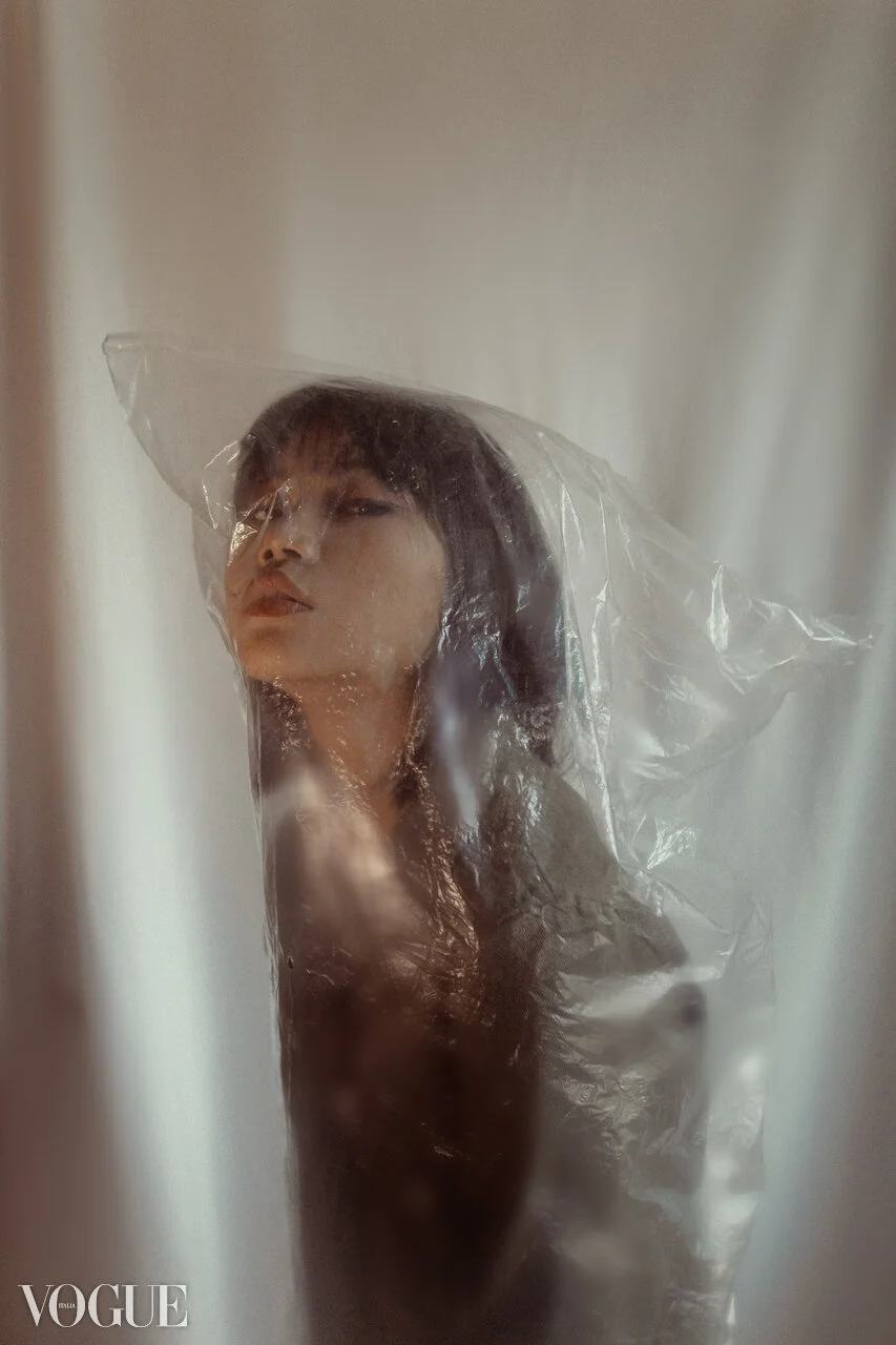 A woman with long dark hair and light makeup, seen through a semi-transparent plastic sheet, with a Vogue magazine logo at the bottom left corner.