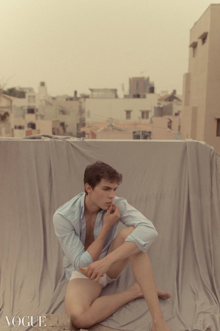 A young man sits on the ground in front of a beige backdrop on a balcony, wearing a partially unbuttoned light blue shirt and white underwear, with a cityscape in the background.