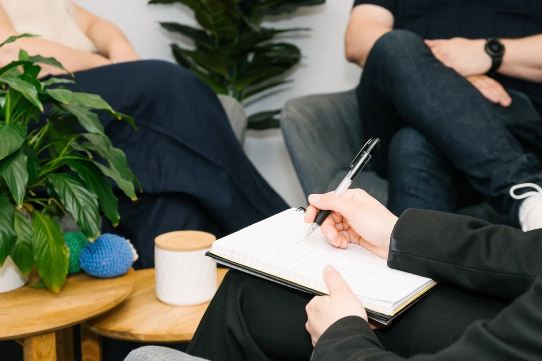 A person taking notes in a notebook during a meeting or therapy session, with two other people sitting nearby and a large green plant on a wooden table.