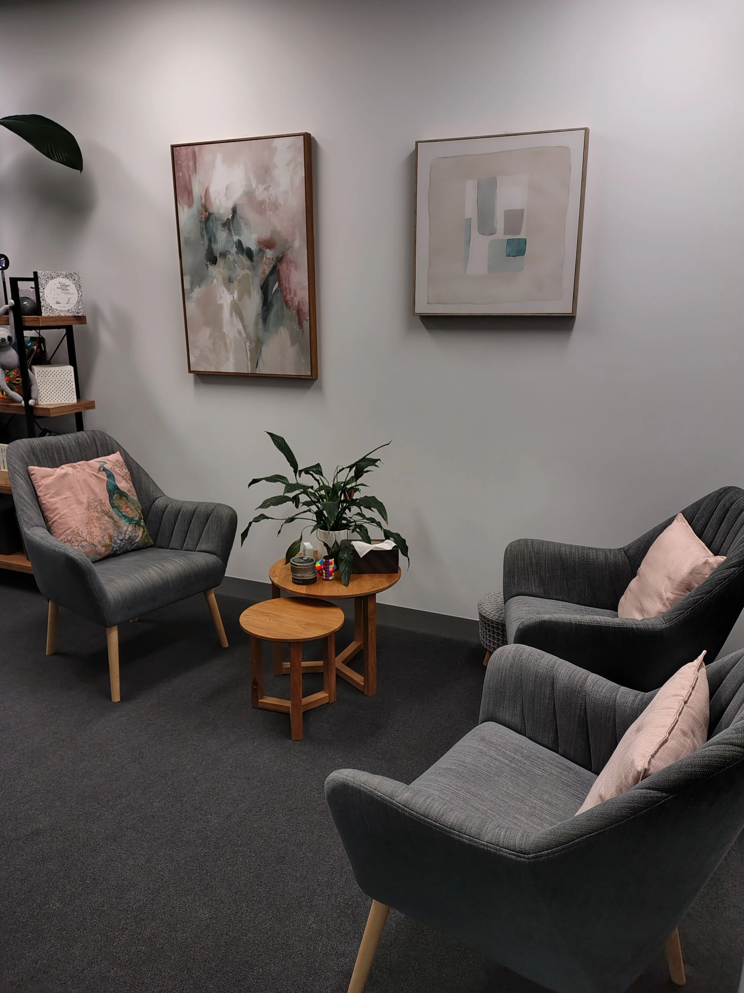 Two gray armchairs with pink pillows and a small wooden round table between them in a room with abstract art on the wall and a black carpet.