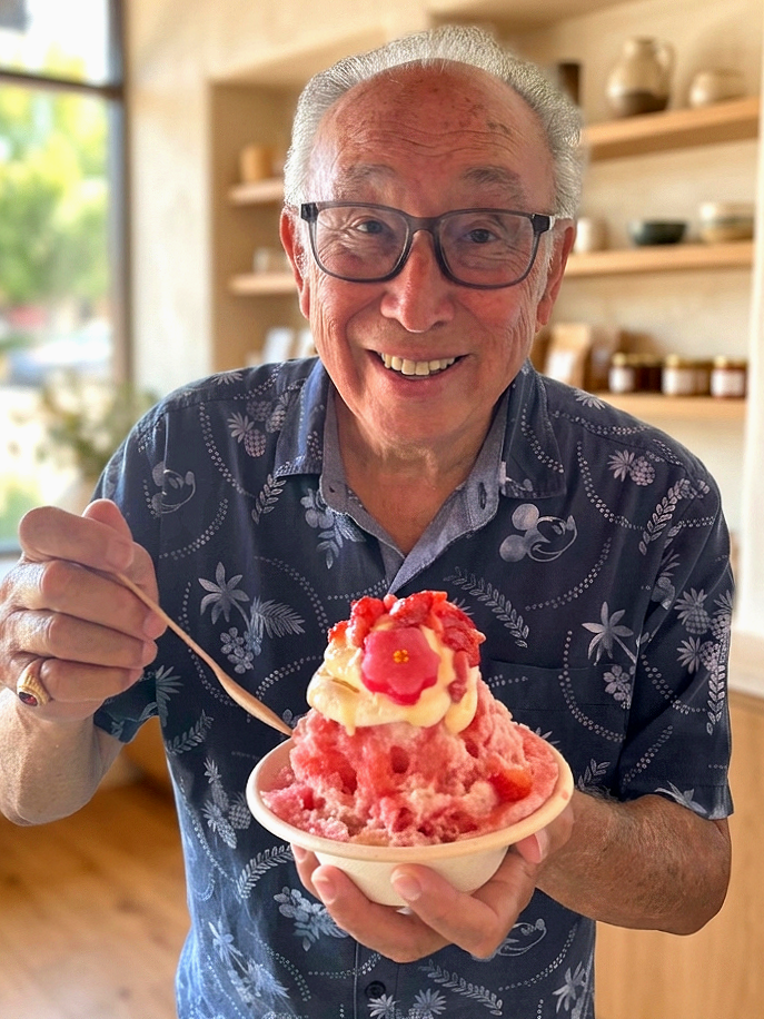 An elderly man with glasses smiling and holding a bowl of pink shaved ice topped with cream and a red flower-shaped topping inside a cozy cafe or bakery.