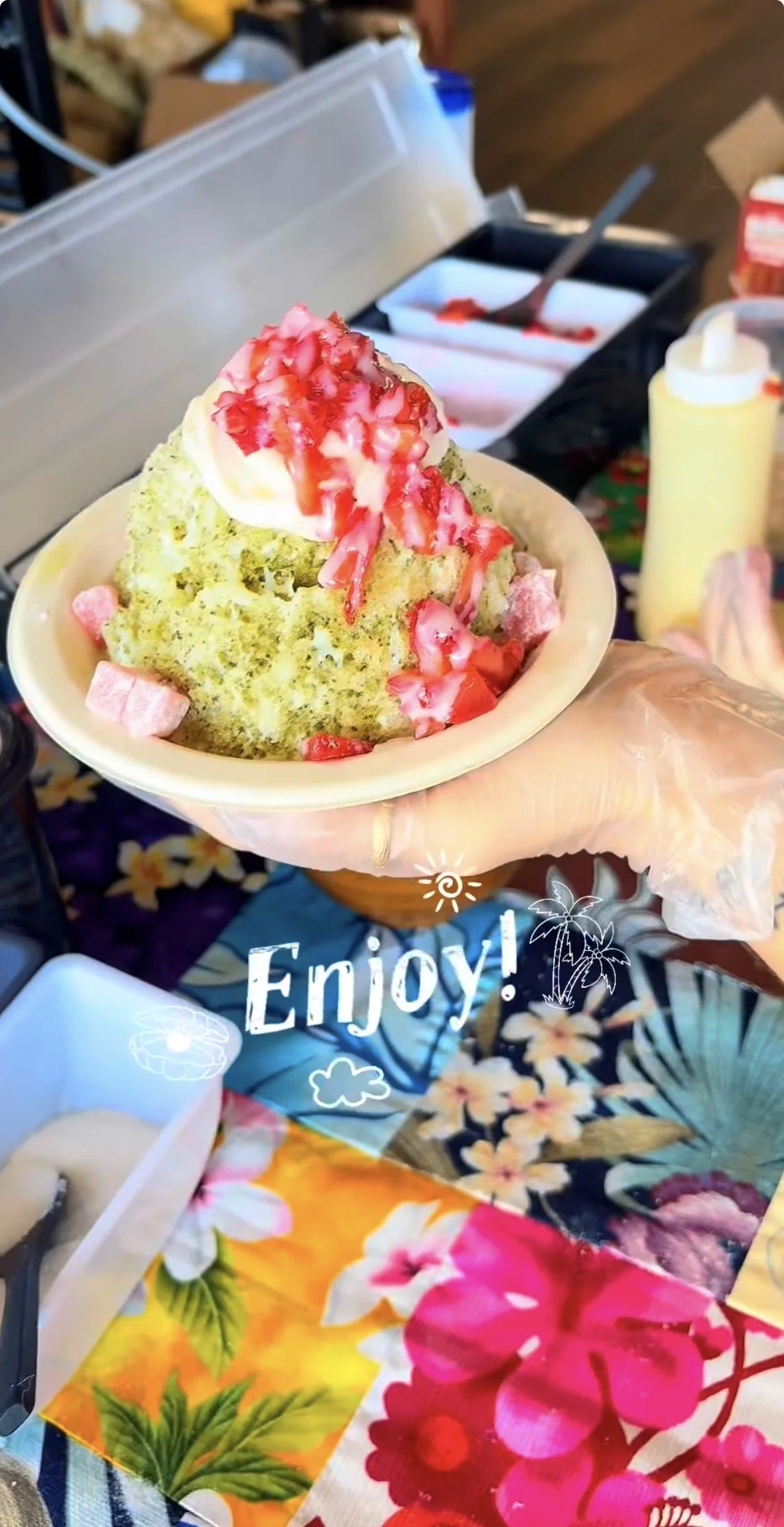 A bowl of shaved ice topped with strawberries and strawberry syrup, held by a person wearing a plastic glove. The background shows a table with condiments and colorful floral decorations with the word "Enjoy!" and emojis.