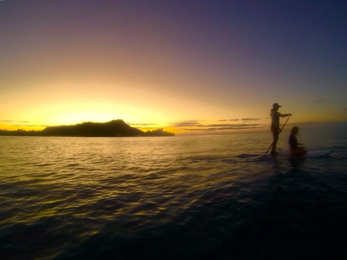 A person and a child riding a paddleboard on the water during sunset, with a mountainous island in the background and the sky filled with purple and yellow hues.