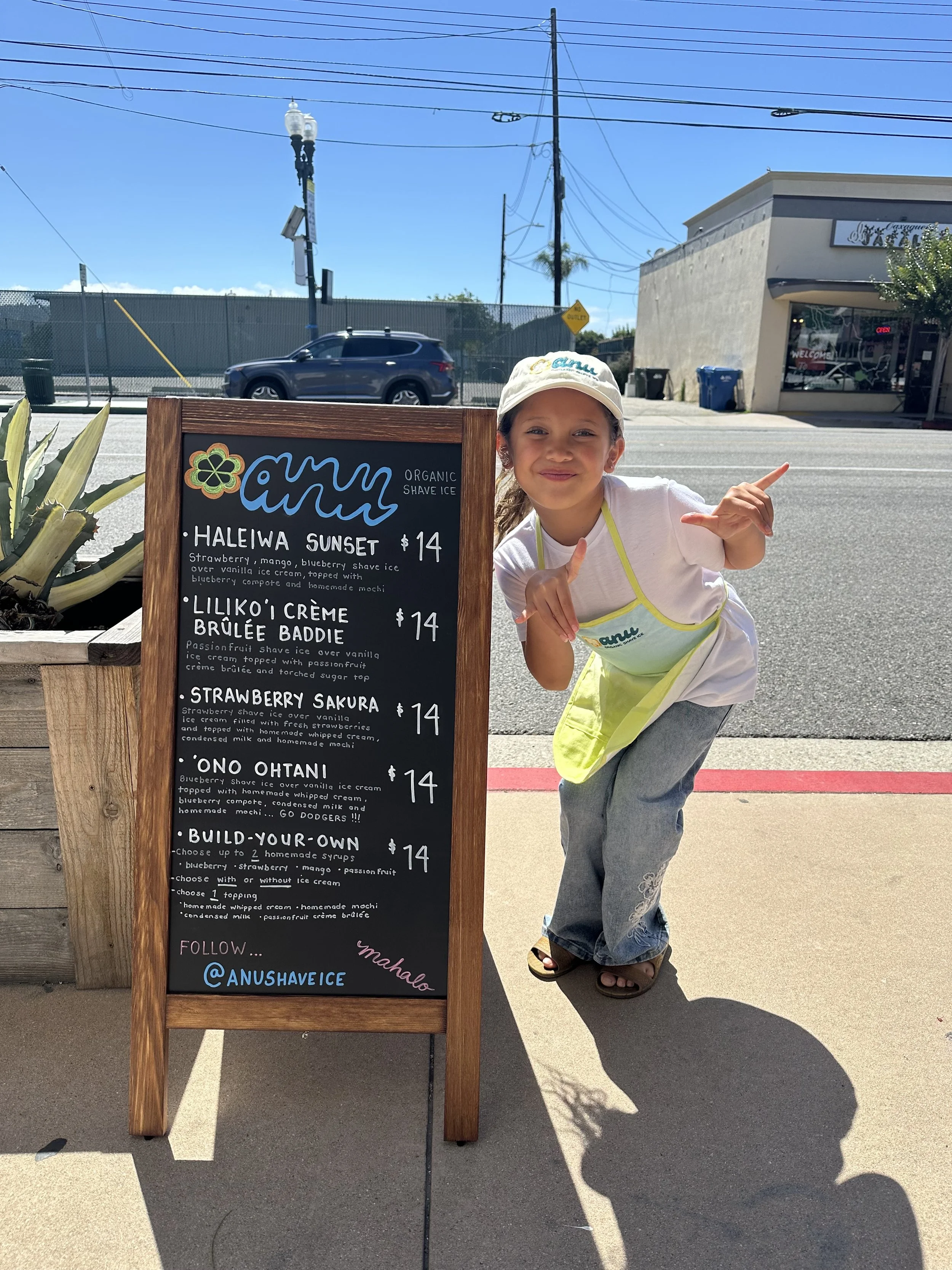 A young girl wearing a white cap, white T-shirt, gray sweatpants with embroidery, and flip-flops, posing beside a blackboard menu for an ice cream shop called 'any Organic Shave Ice.' The menu lists various shave ice flavors and prices, with a bright, sunny outdoor setting, a parking lot, a building with a shopfront, and a few trees in the background.