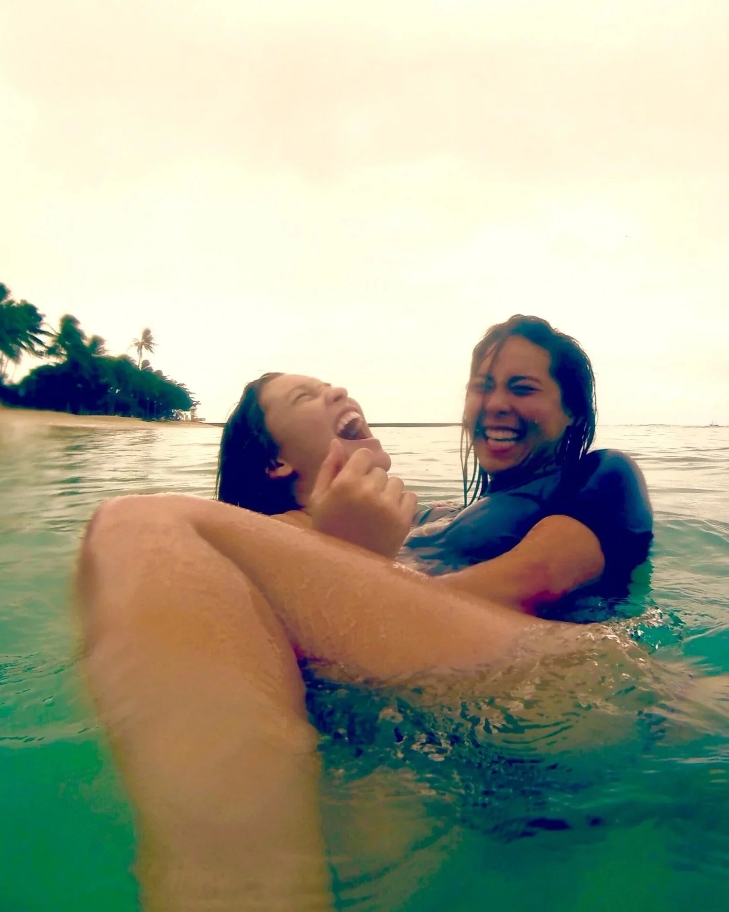 Two smiling women laughing and enjoying themselves while swimming in the ocean near a tropical island with palm trees under a bright sky.