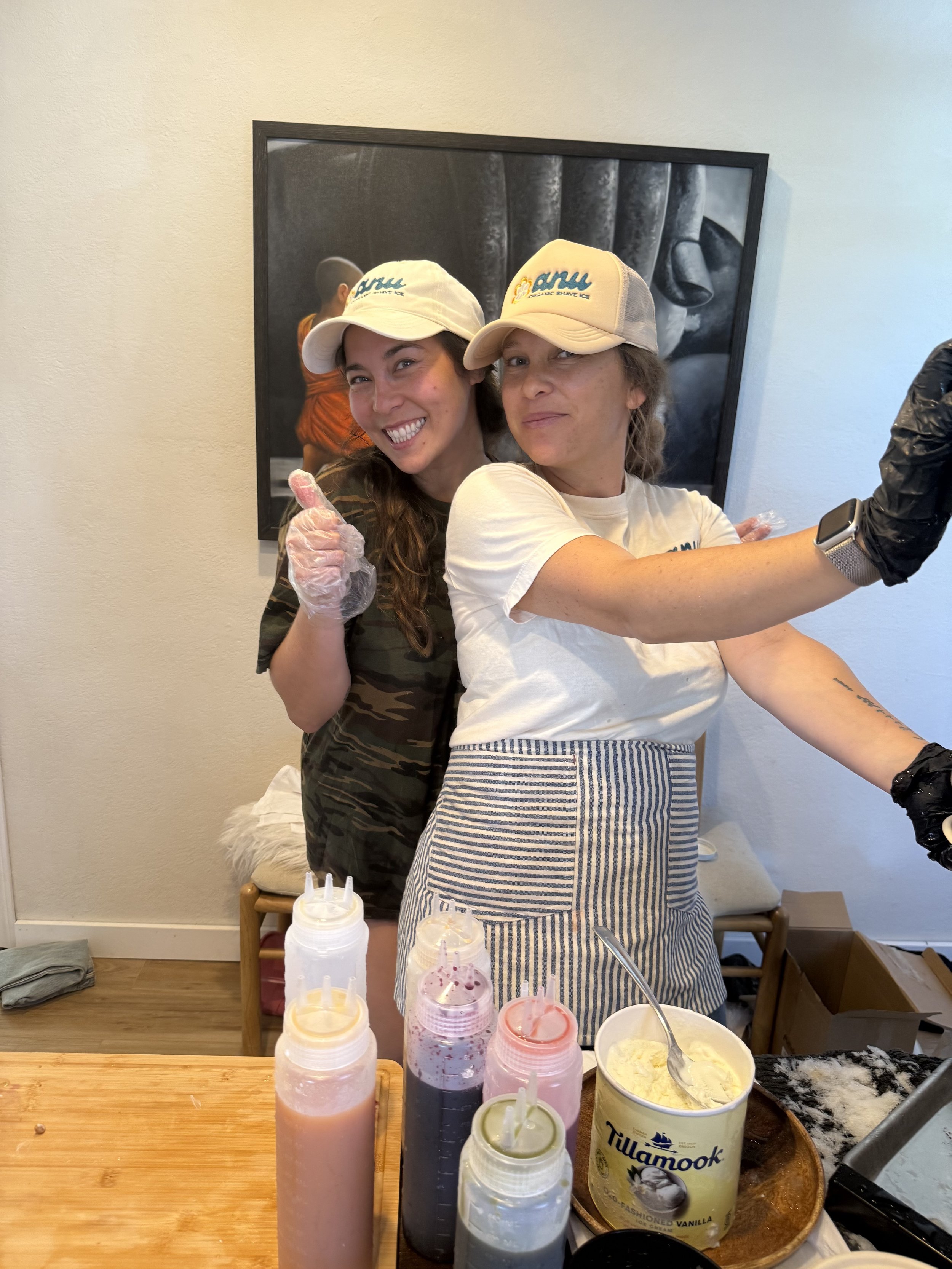 Two women wearing beige caps and aprons, preparing ice cream or dessert with syrups and toppings in a kitchen or café setting.