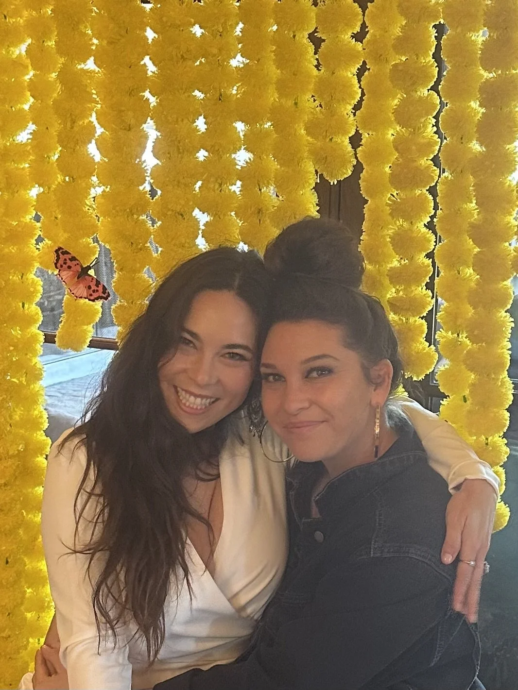 Two women smiling and embracing in front of a backdrop of yellow marigold flowers and hanging yellow marigolds with a pink butterfly decoration.