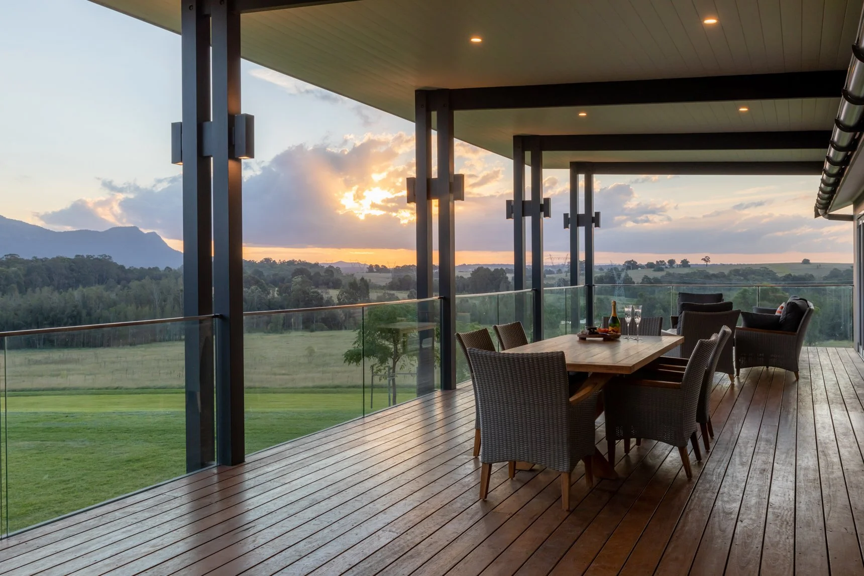 Outdoor balcony with wooden flooring and glass railing, set with a dining table, chairs, and a seating area, overlooking a scenic landscape of trees, fields, and mountains at sunset.