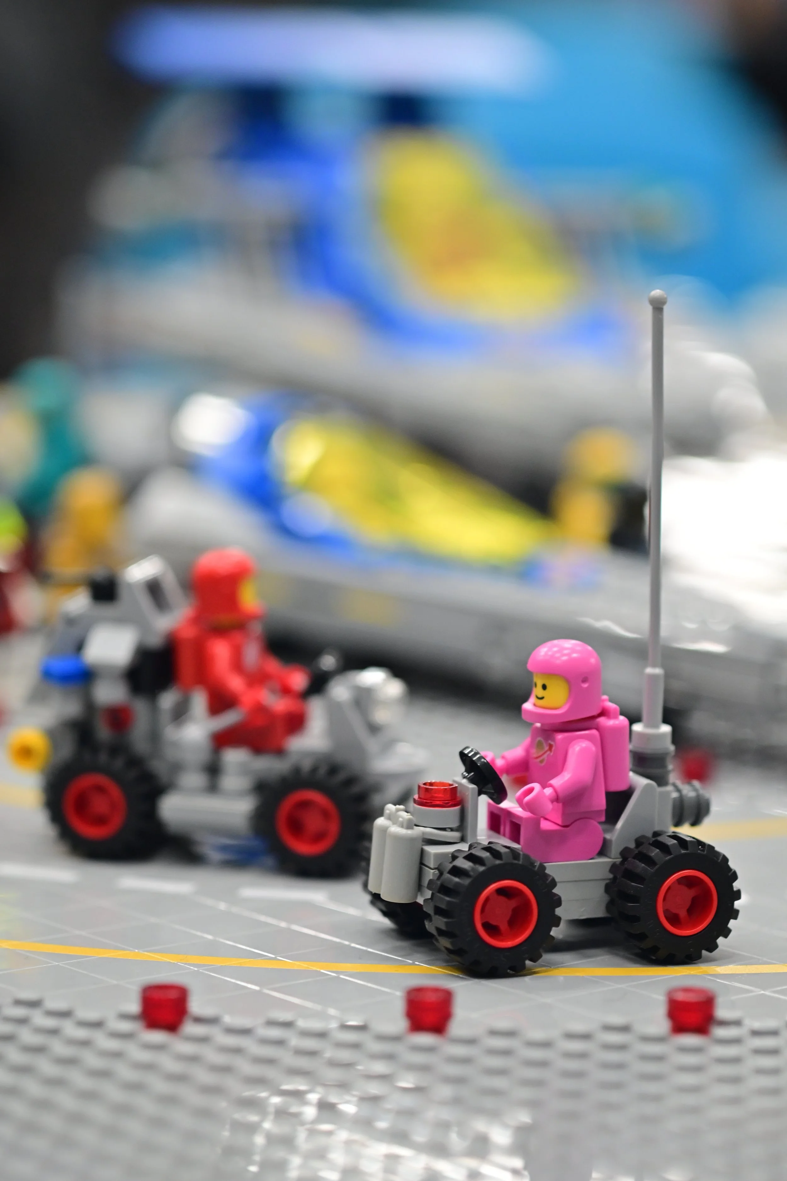 LEGO space-themed scene with a pink astronaut minifigure driving a small white rover with black wheels, a red control panel, and a long antenna. The background features blurred colorful elements and a gray surface with yellow lines, mimicking a plane
