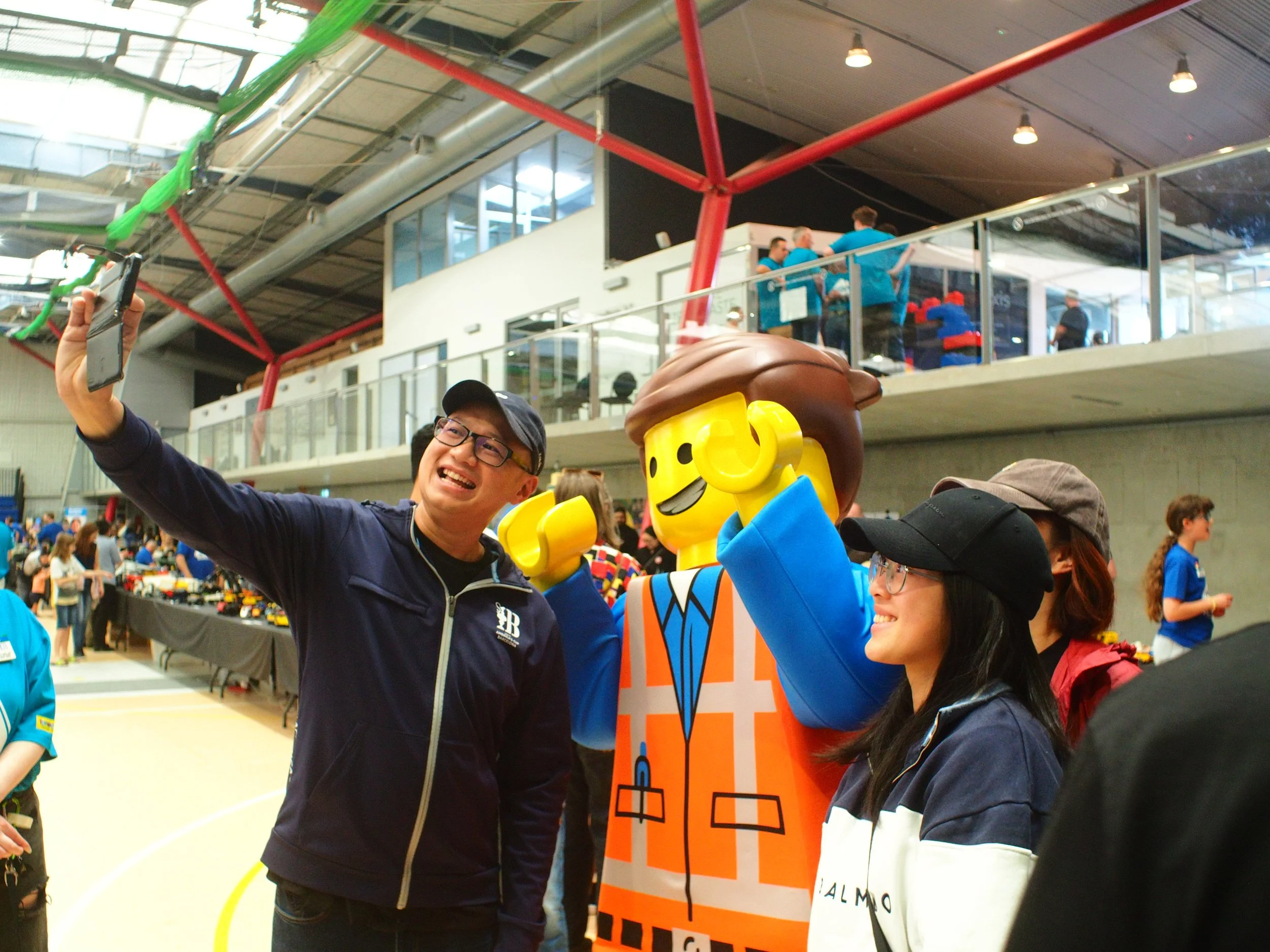 People celebrating at an indoor event with a large LEGO character mascot and banners.