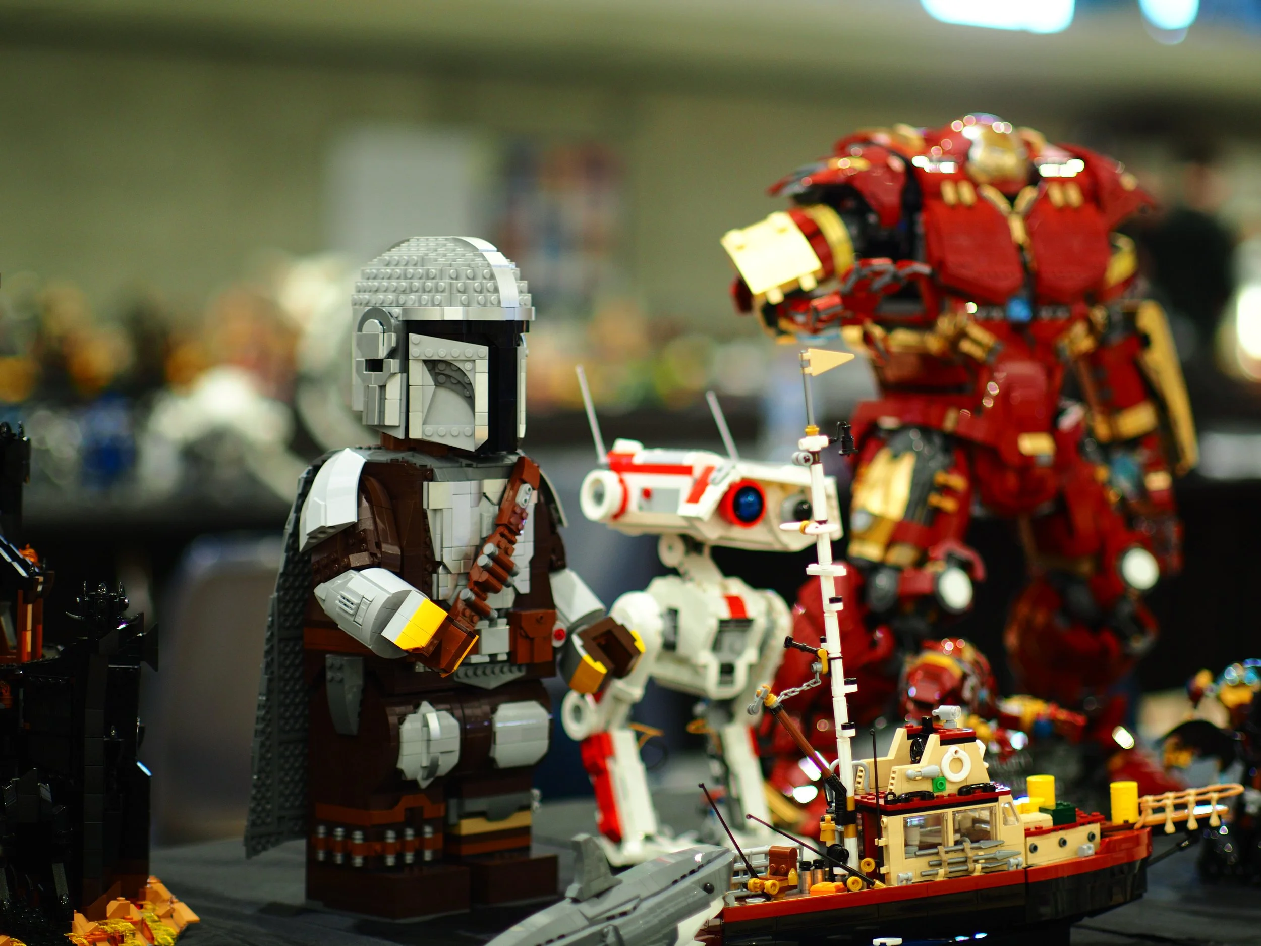 Display of various LEGO model vehicles and characters, including a gray and brown LEGO character in the foreground and larger LEGO vehicles and figures in the background, set up on a table or display surface.