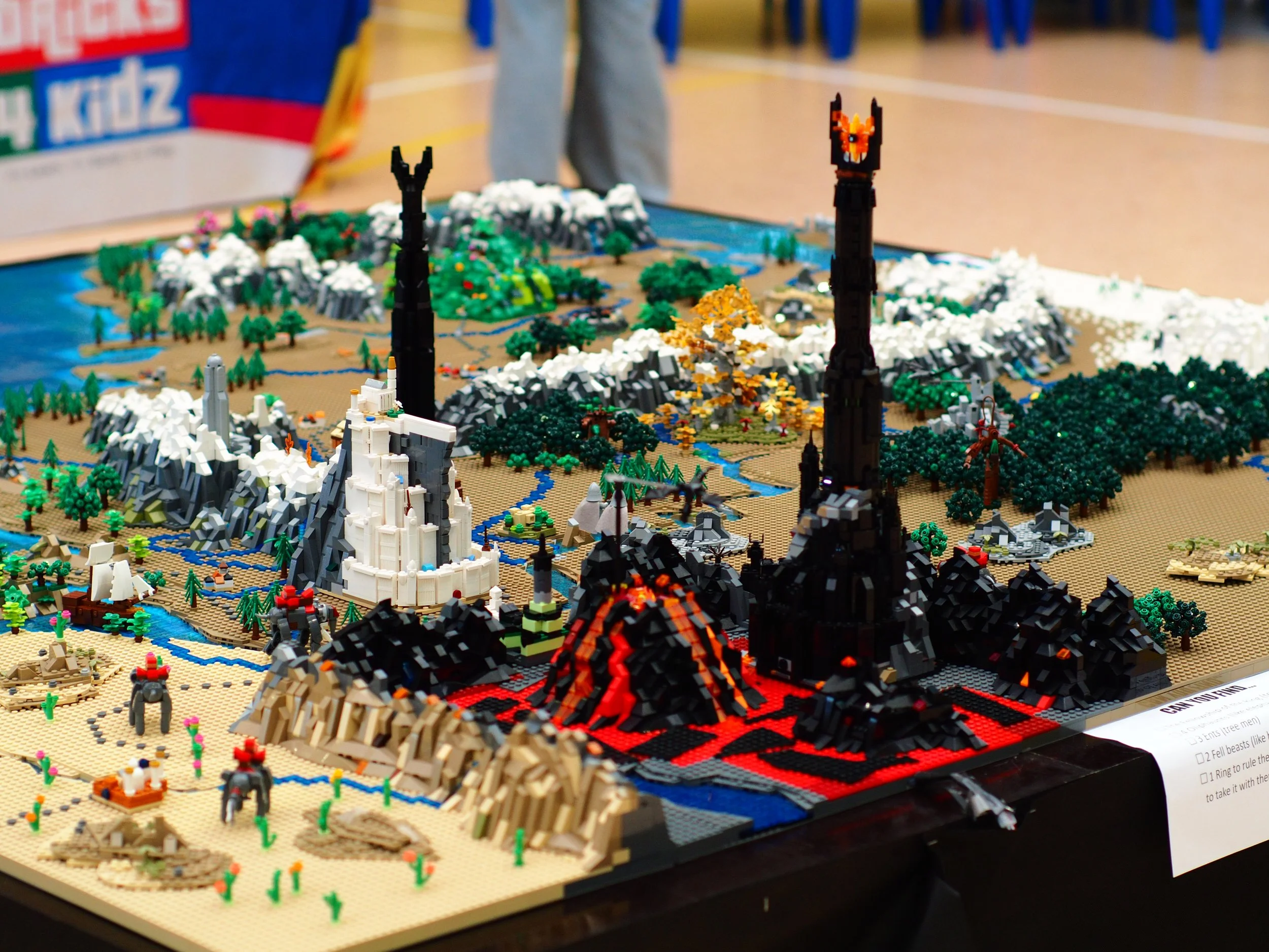LEGO diorama depicting a volcanic eruption with lava flowing down, surrounded by various LEGO trees, mountains, and small structures, on a display table at an exhibition.