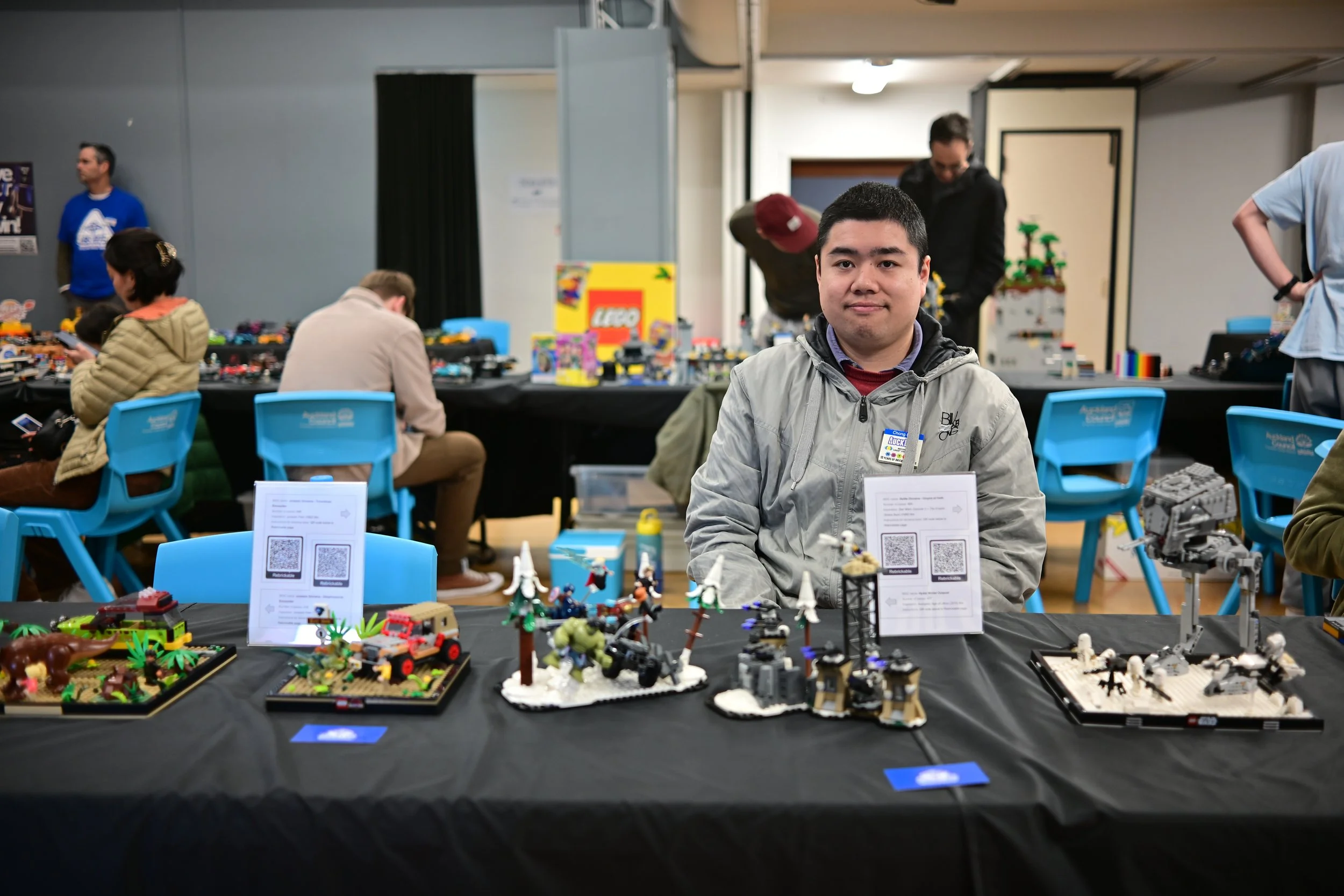 A young man sitting at a table displaying various LEGO models at a LEGO exhibition. He wears a gray jacket and a name tag. In the background, other people are seated at tables with LEGO sets, and some are standing and browsing. The room has gray wall