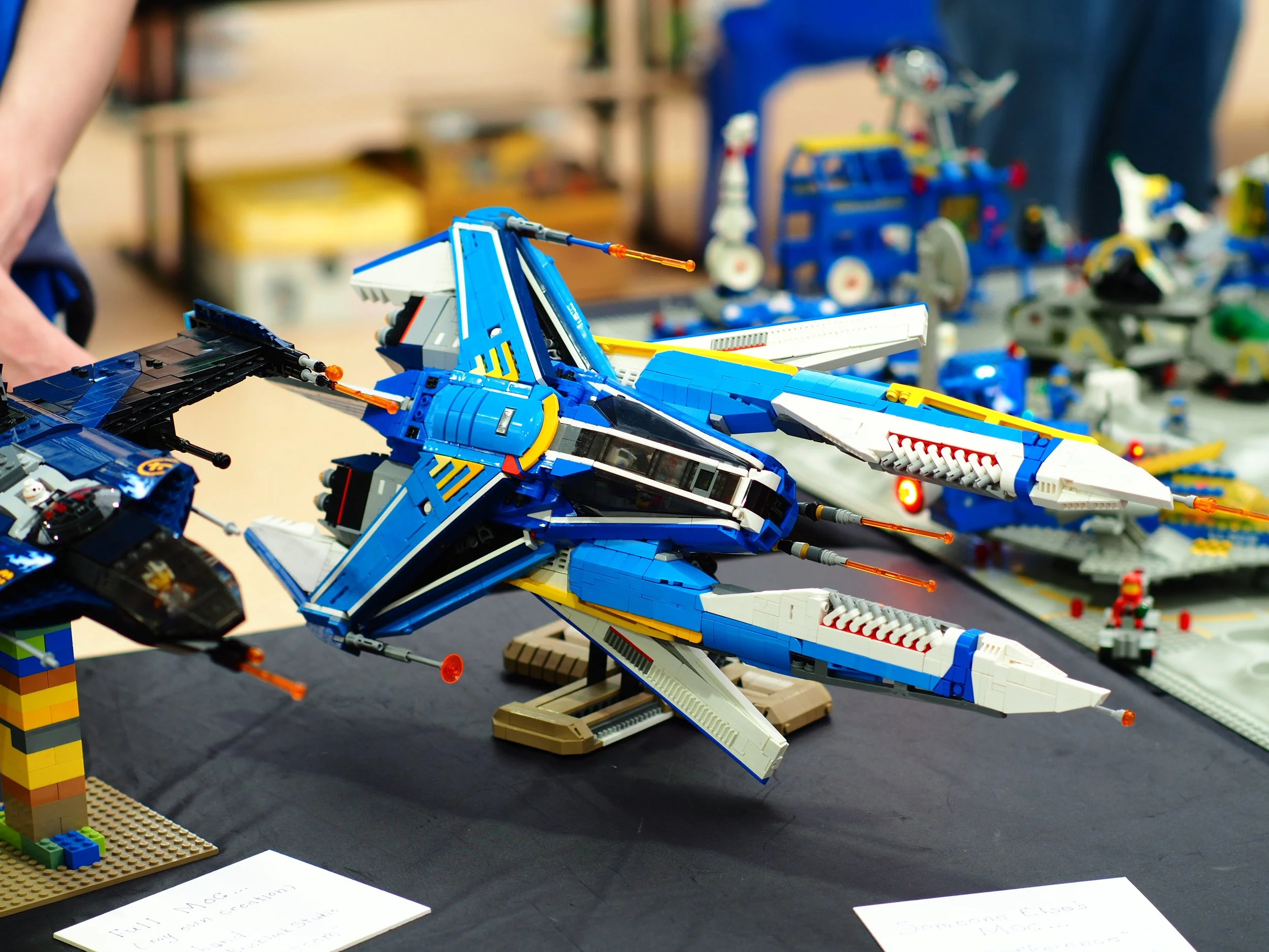 Lego spaceship model blue and white with orange accents on display at a Lego exhibition table with handwritten notes in front.