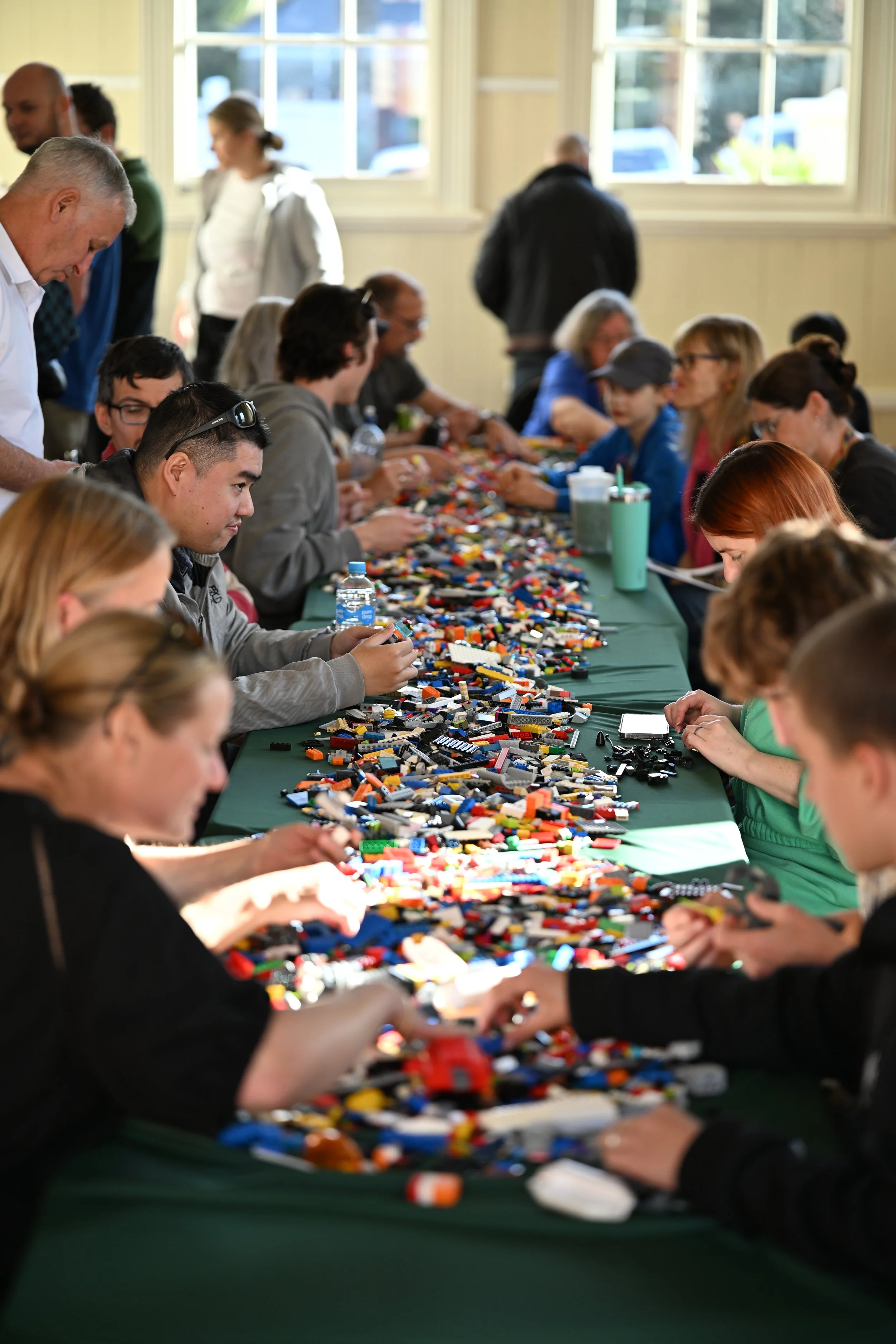 Multiple individuals sitting around a table filled with a large collection of LEGO pieces, engaging in building or sorting. The setting appears to be a community center or event space with large windows and natural light.
